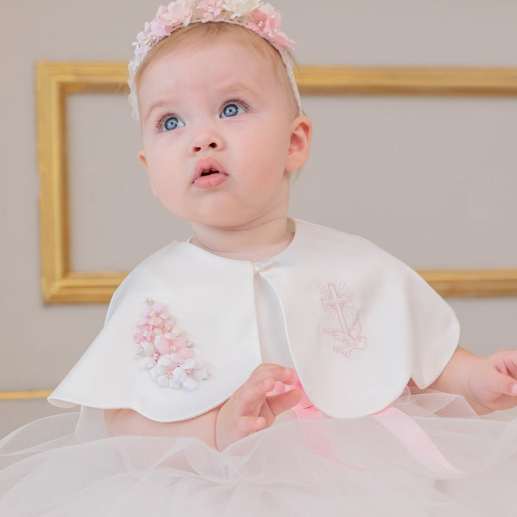 A baby girl wearing a white and pink first birthday dress with a tulle skirt, a white cape with pink floral embroidery, and a matching pink floral headband, sitting on a light wooden floor.