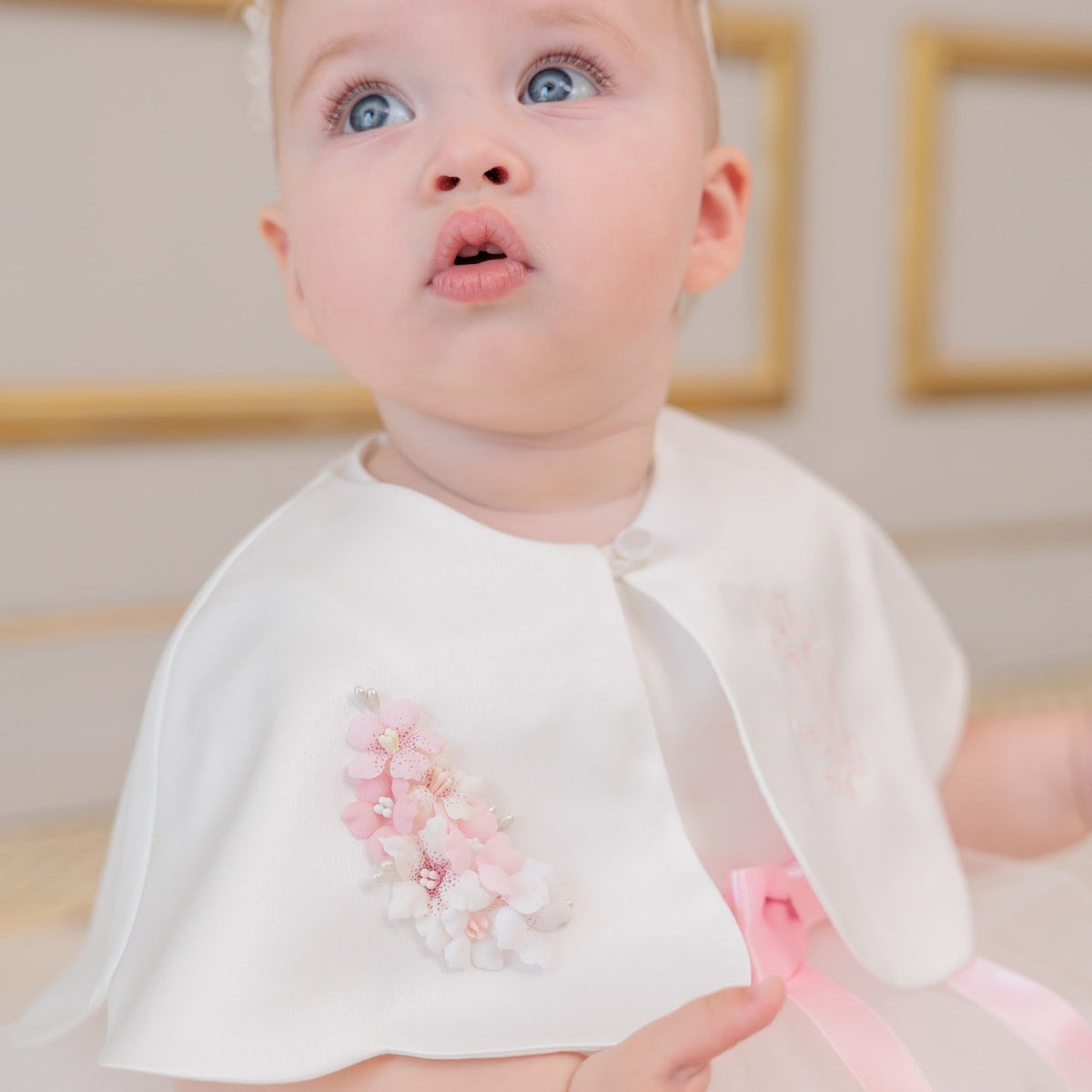 A baby girl wearing a white tulle dress with a matching white cape featuring pink and white floral embroidery, a pink ribbon, and a pink floral headband.
