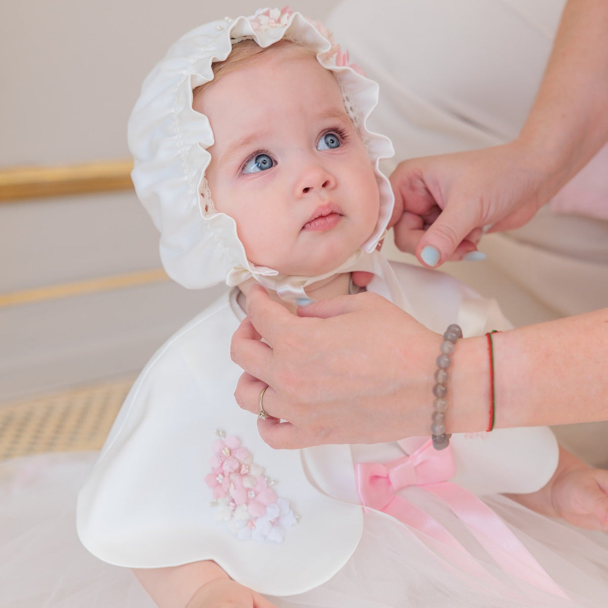 A baby wearing a white bonnet and white dress with pink ribbon details, while an adult's hands adjust the bonnet. The background is a soft beige indoor setting.