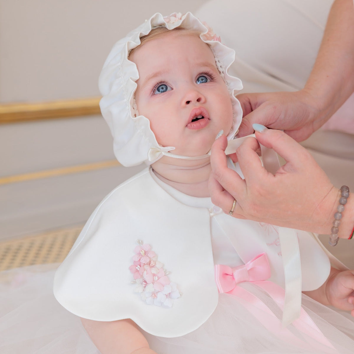 A baby wearing a white bonnet and white tulle dress with pink ribbon accents, seated while an adult adjusts the bonnet. The scene is softly lit and styled indoors.