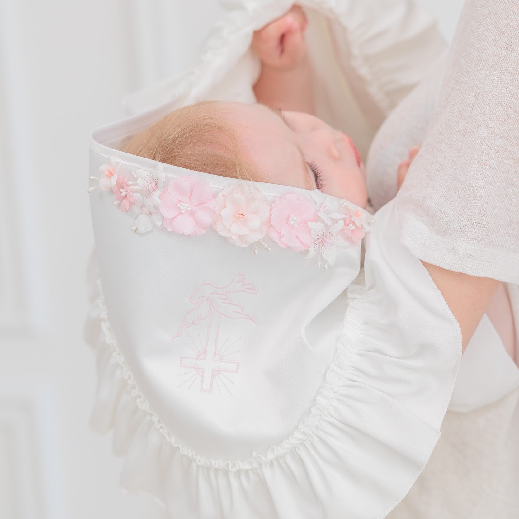 A close-up lifestyle shot of a baby wearing a white satin bonnet with pink floral trim and delicate embroidery, held by an adult in a matching white outfit against a bright, soft background.