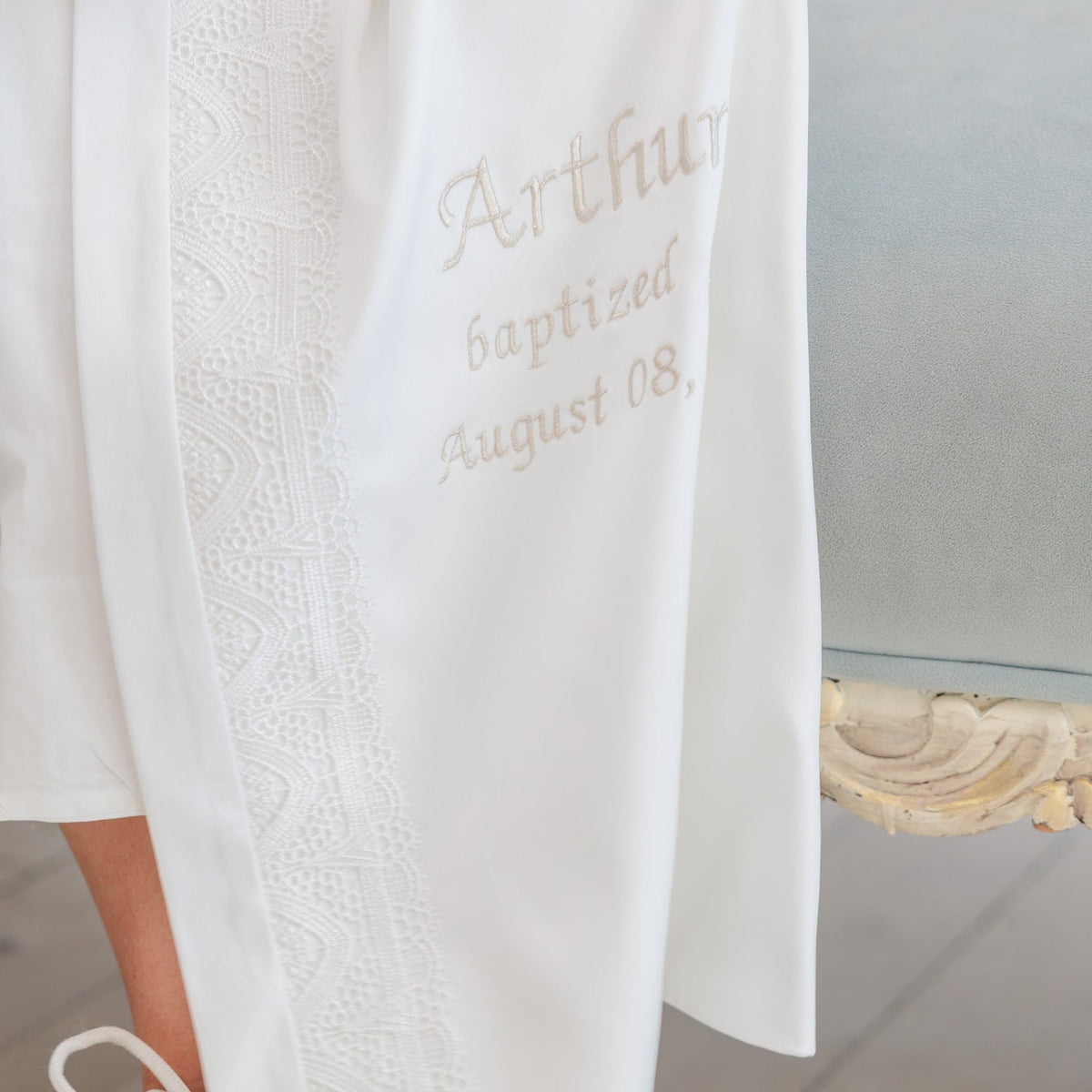 Close-up of a white embroidered robe with lace trim and the name 'Arthur' and date 'August 08' embroidered on the front, worn by a person seated indoors near a light-colored chair.