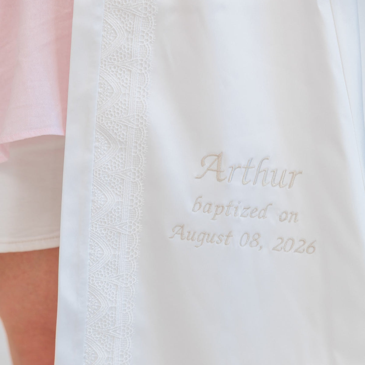Close-up of a white baptismal gown or ceremonial garment with embroidered text reading 'Arthur baptized on August 08, 2026' and a decorative lace-like border, worn by a person whose legs are visible.