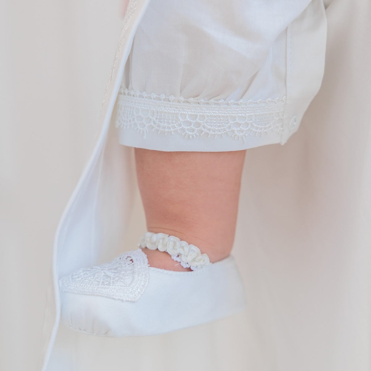 A close-up of a baby's foot wearing a white lace-trimmed bootie, tucked under a matching white robe.