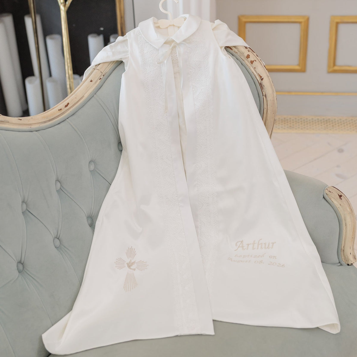 A white long-sleeve baby cape or smock with delicate embroidery, hanging on a hanger from an ornate light blue tufted sofa in a luxurious room.