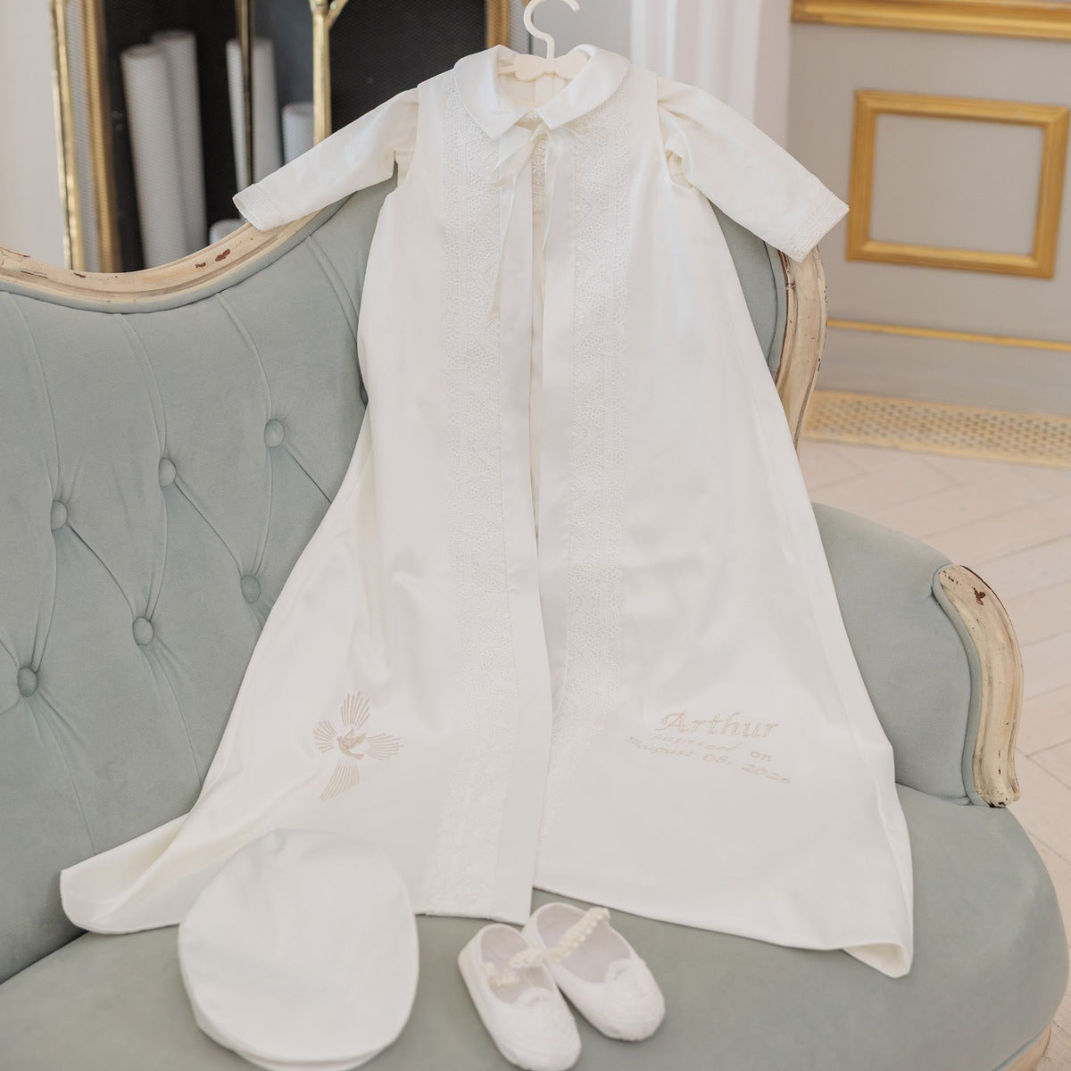 A white baby christening gown with short sleeves and a collar, displayed on a hanger on a tufted light blue-green sofa. A matching white bonnet and white satin booties are placed in front.