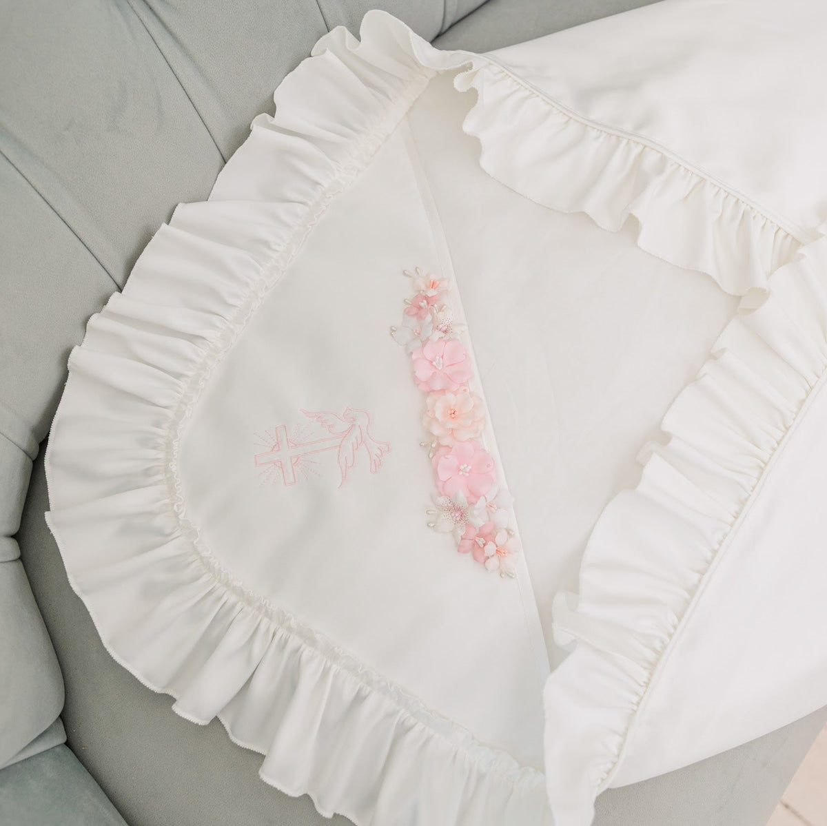 A white ruffled baby blanket or pillow cover with delicate pink floral embroidery, styled on a light gray tufted velvet sofa.