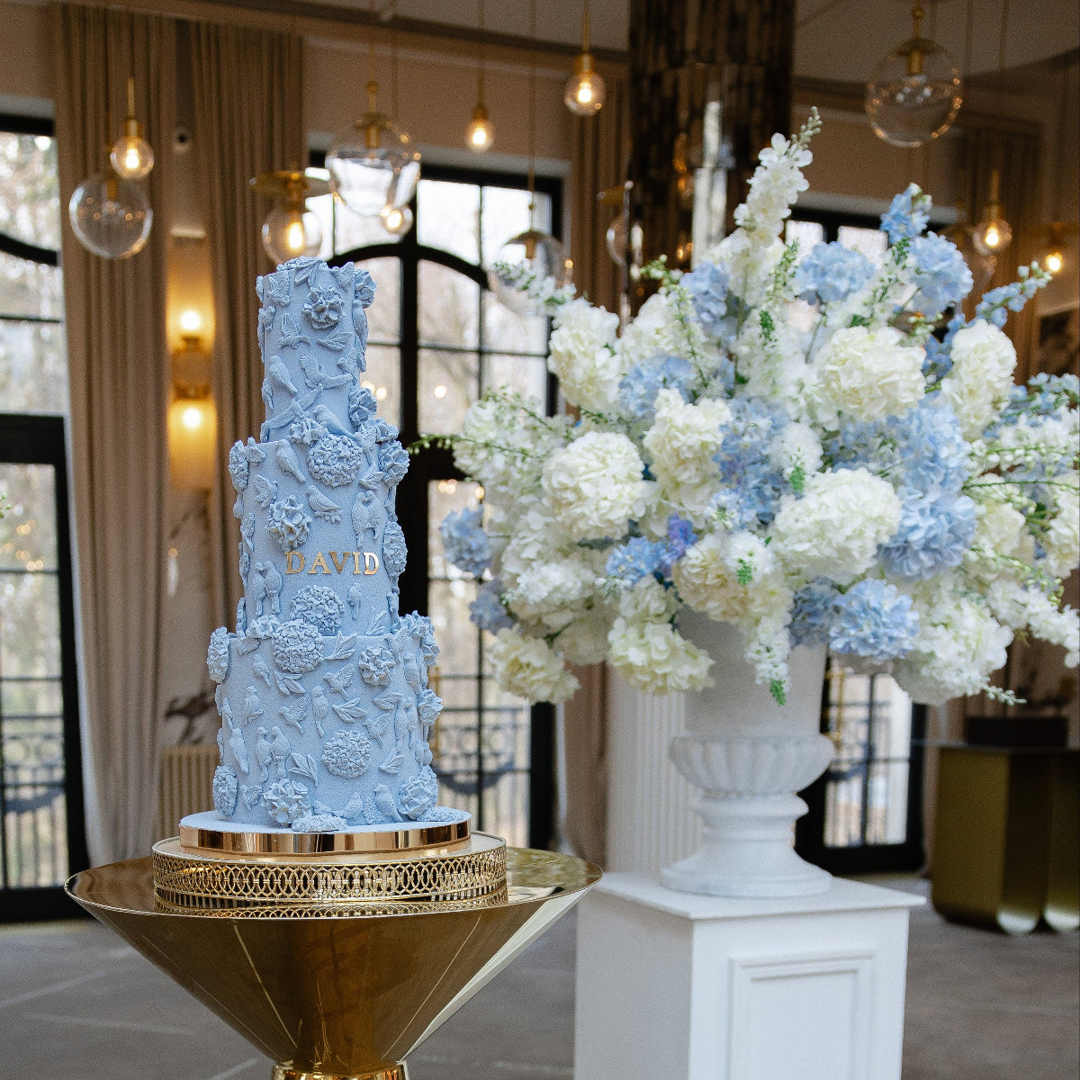 Decorative blue baptism boy cake on a gold stand with floral arrangements in an indoor setting.