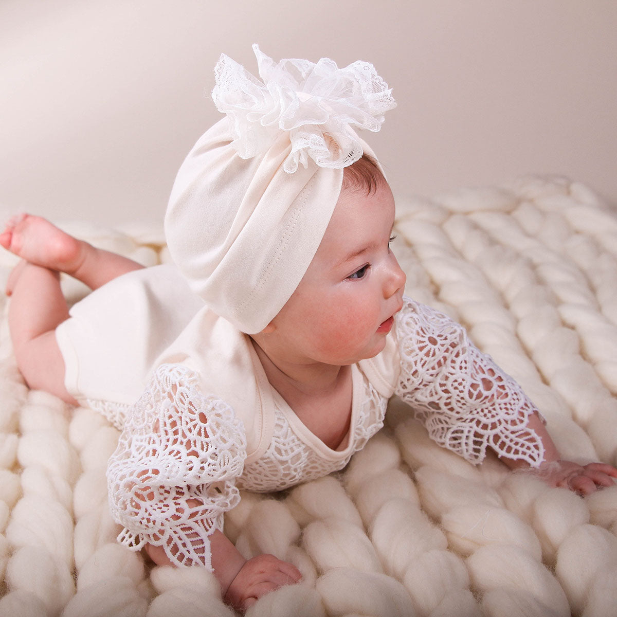 Baby wearing a white lace outfit and headband on a soft, textured surface
