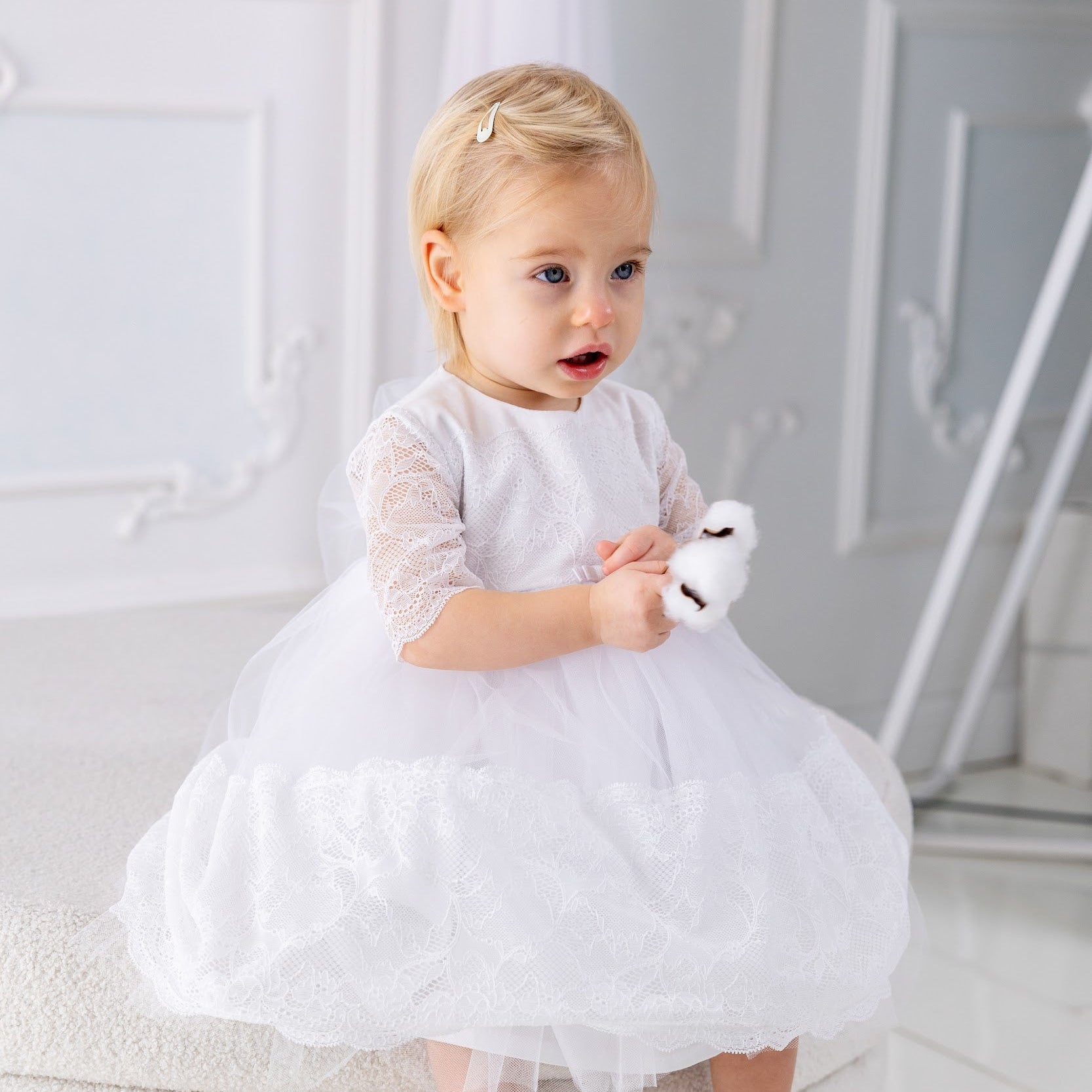 Baby in a white baptism dress sitting on a white cushion holding a white teddy bear.