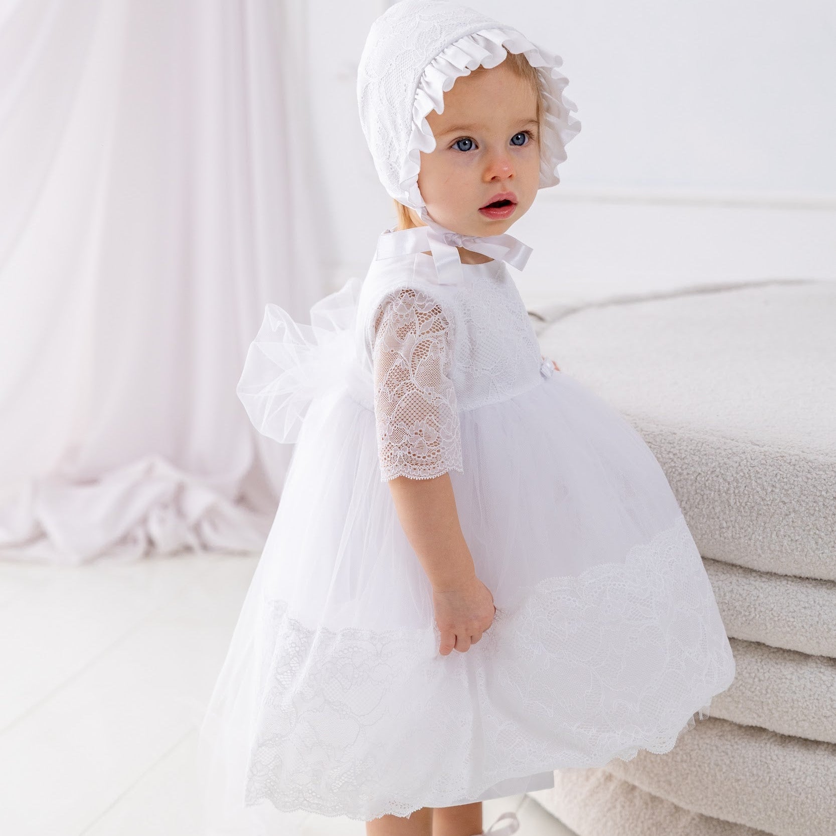 Baby in a white dress and bonnet standing on a light wooden floor.