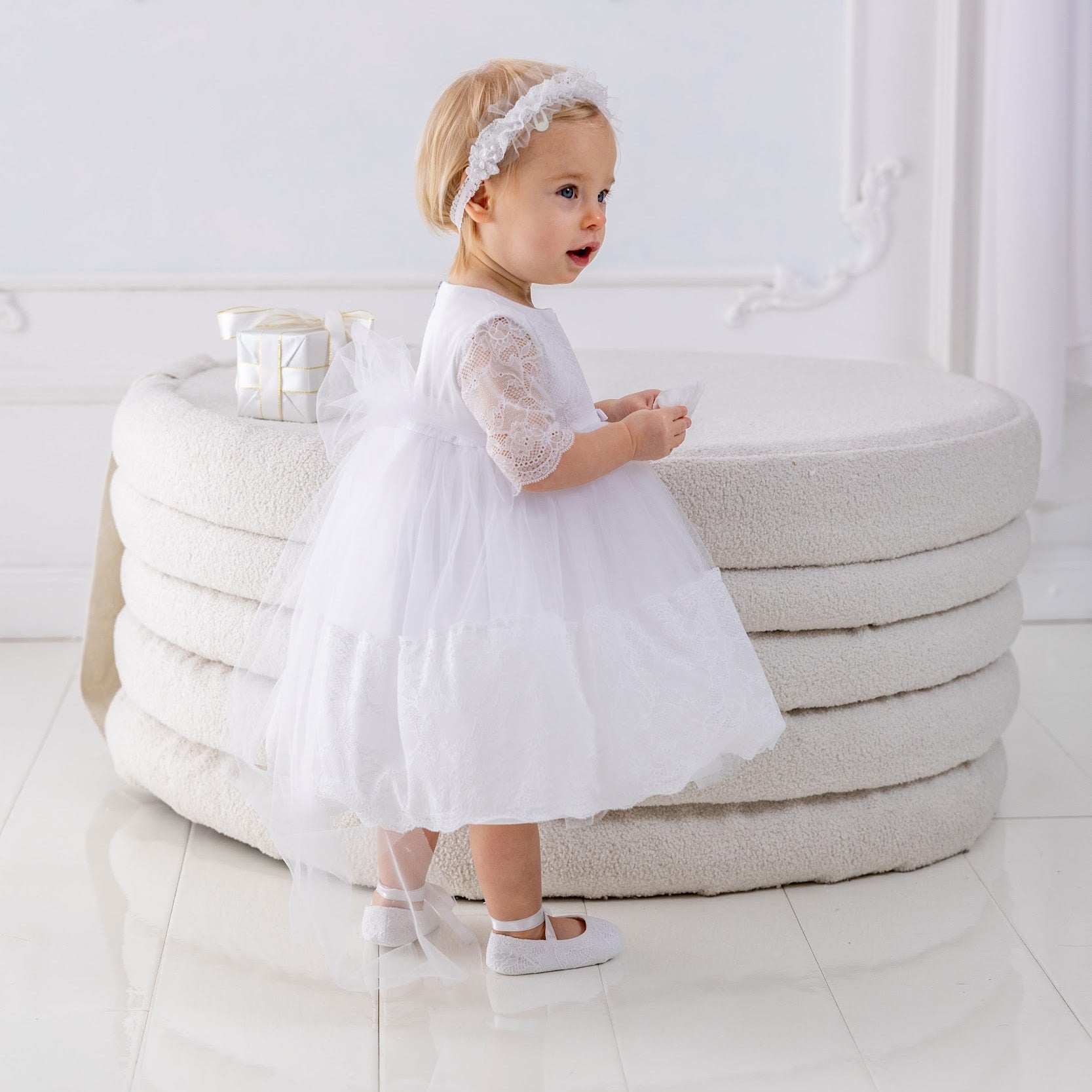 Young child in a white dress standing on a white floor with a decorative white wall in the background.