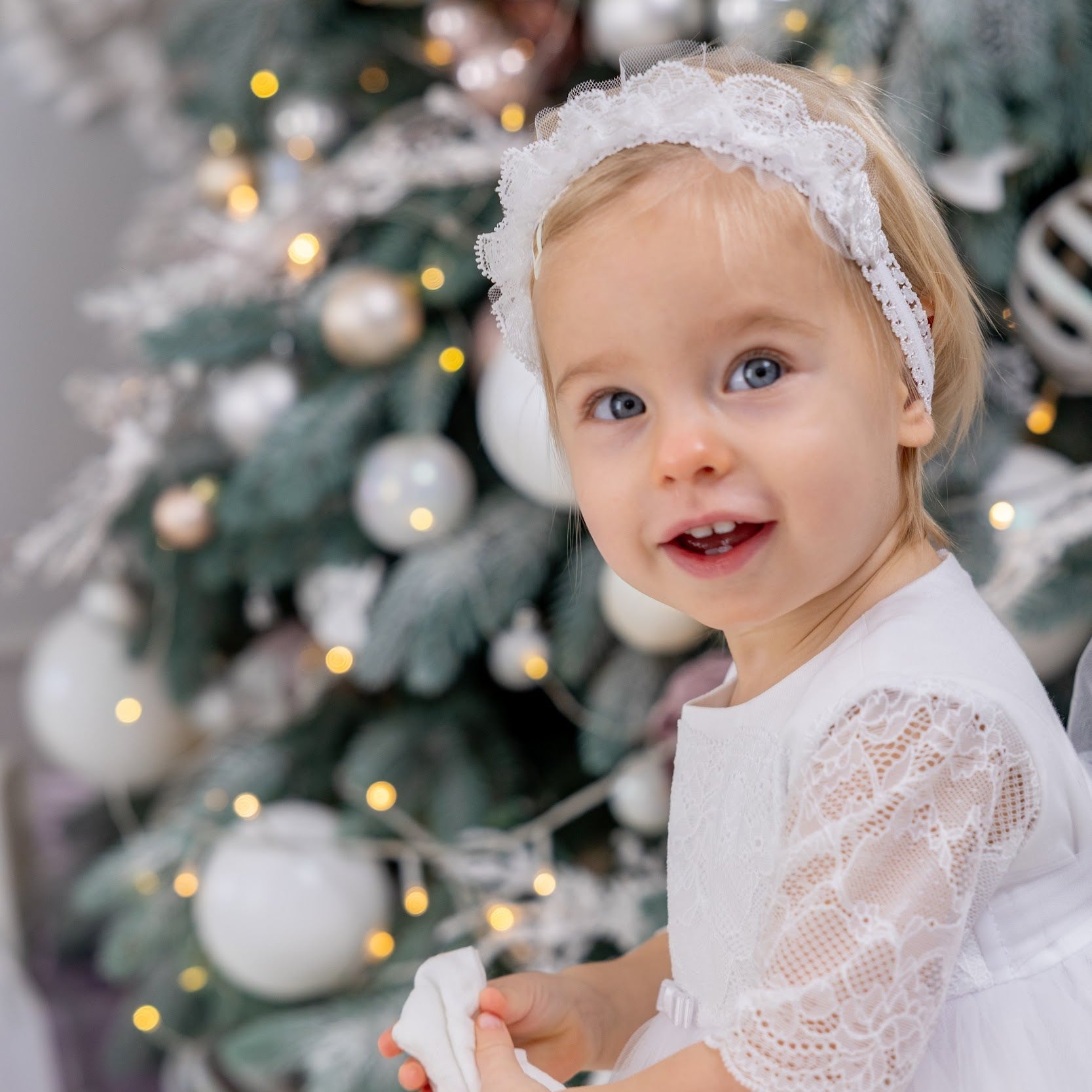 Child in a white dress standing in front of a decorated Christmas tree.