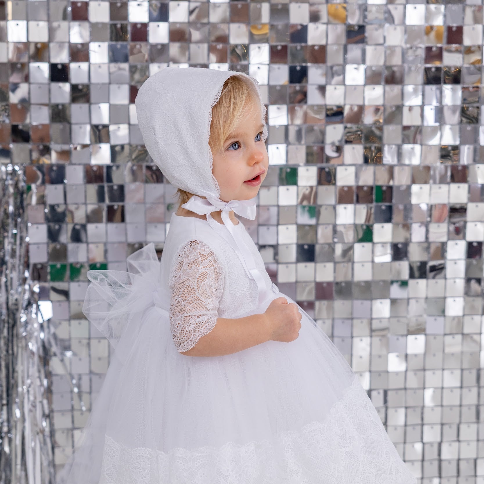Child in a white dress with a hood standing against a reflective silver wall.