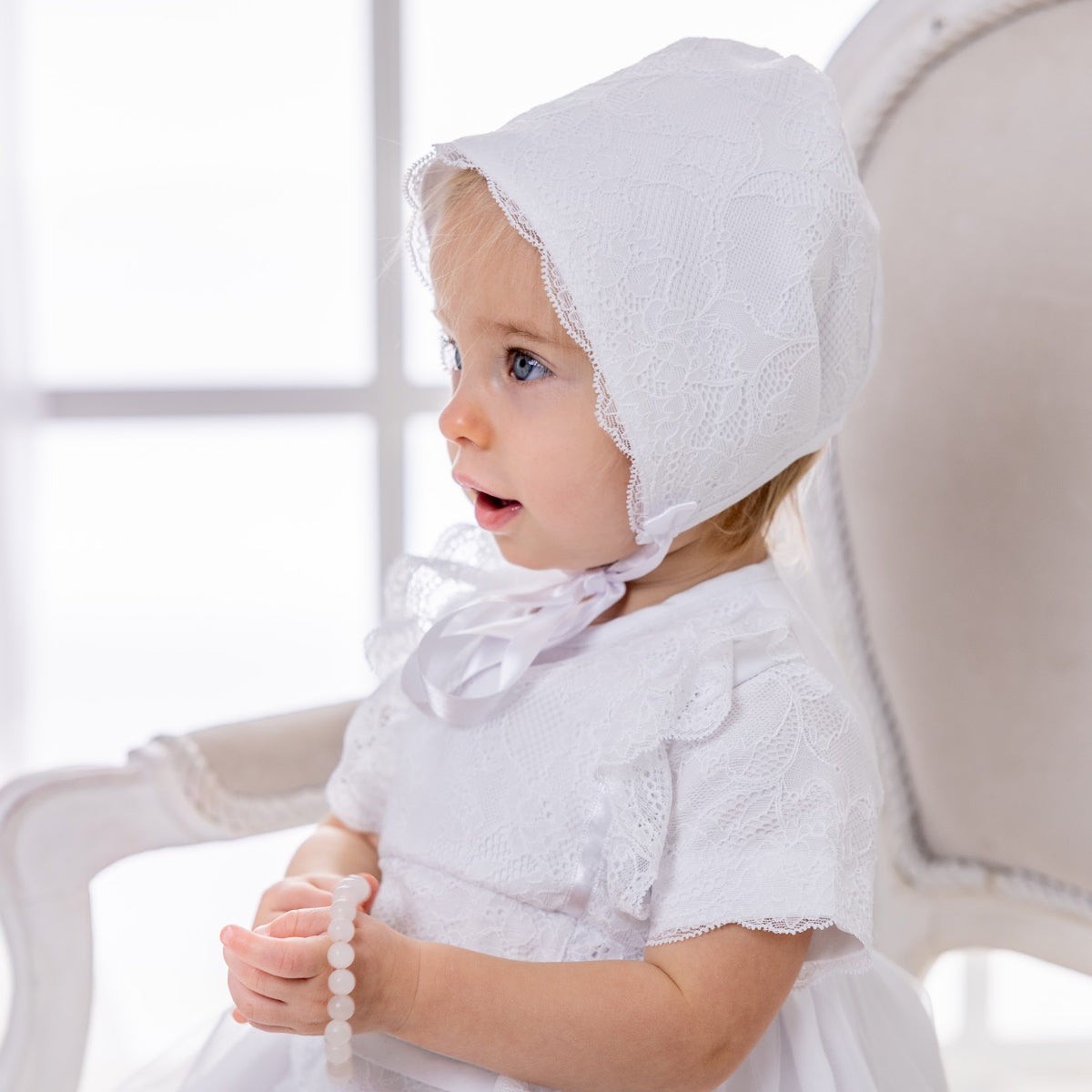 Baby wearing a white lace bonnet sitting on a chair.