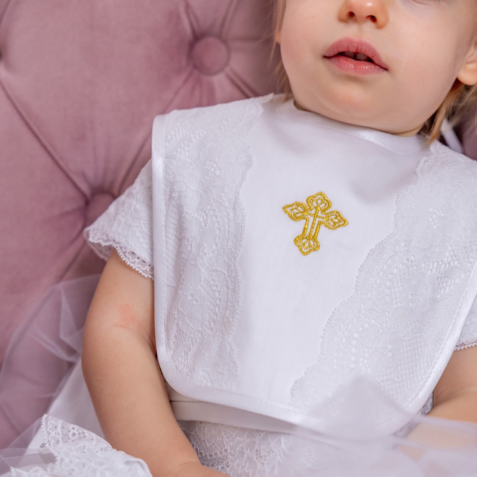 Child in a white dress with a gold cross detail, sitting on a pink cushion.