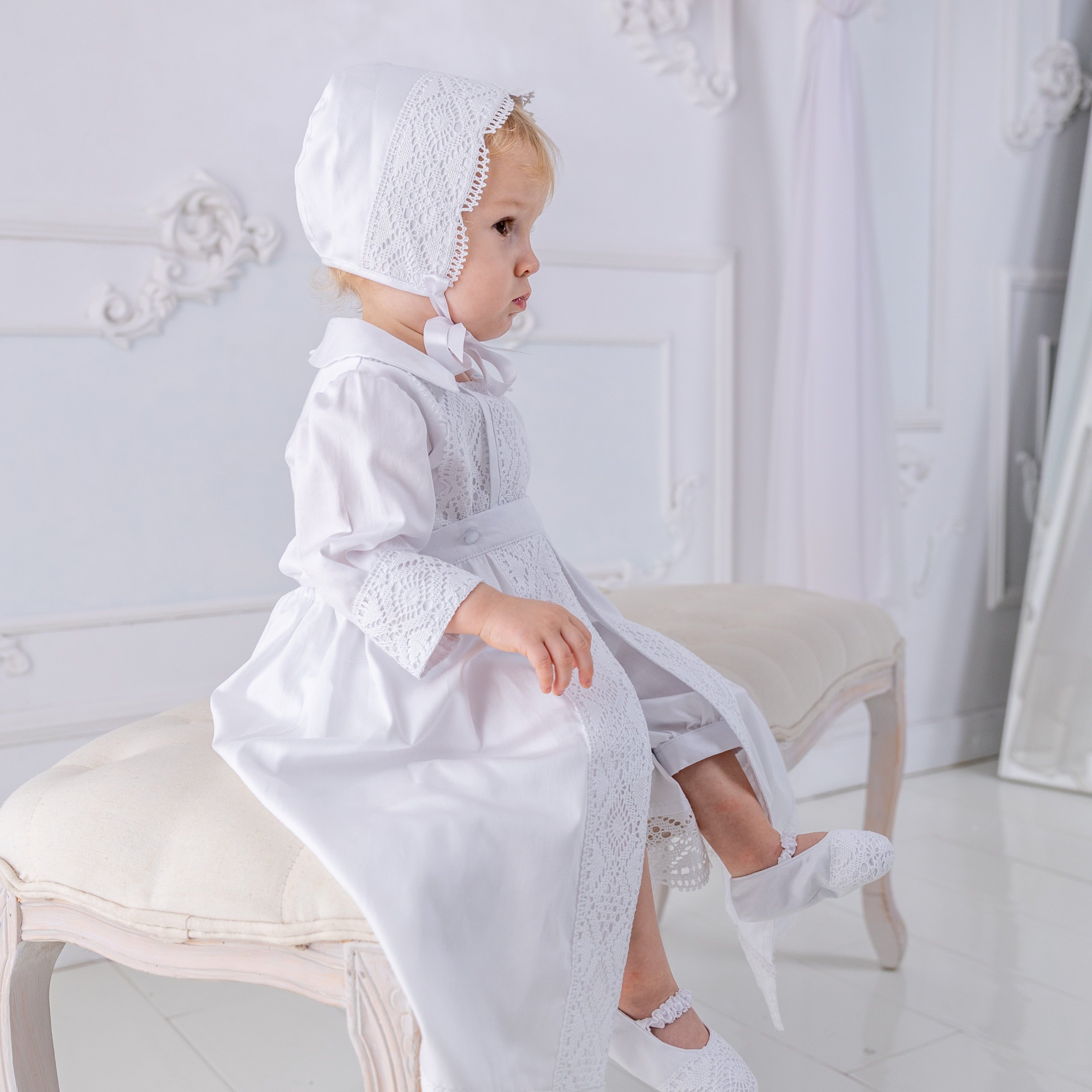 Baby in a white outfit sitting on a stool in a decorated room.