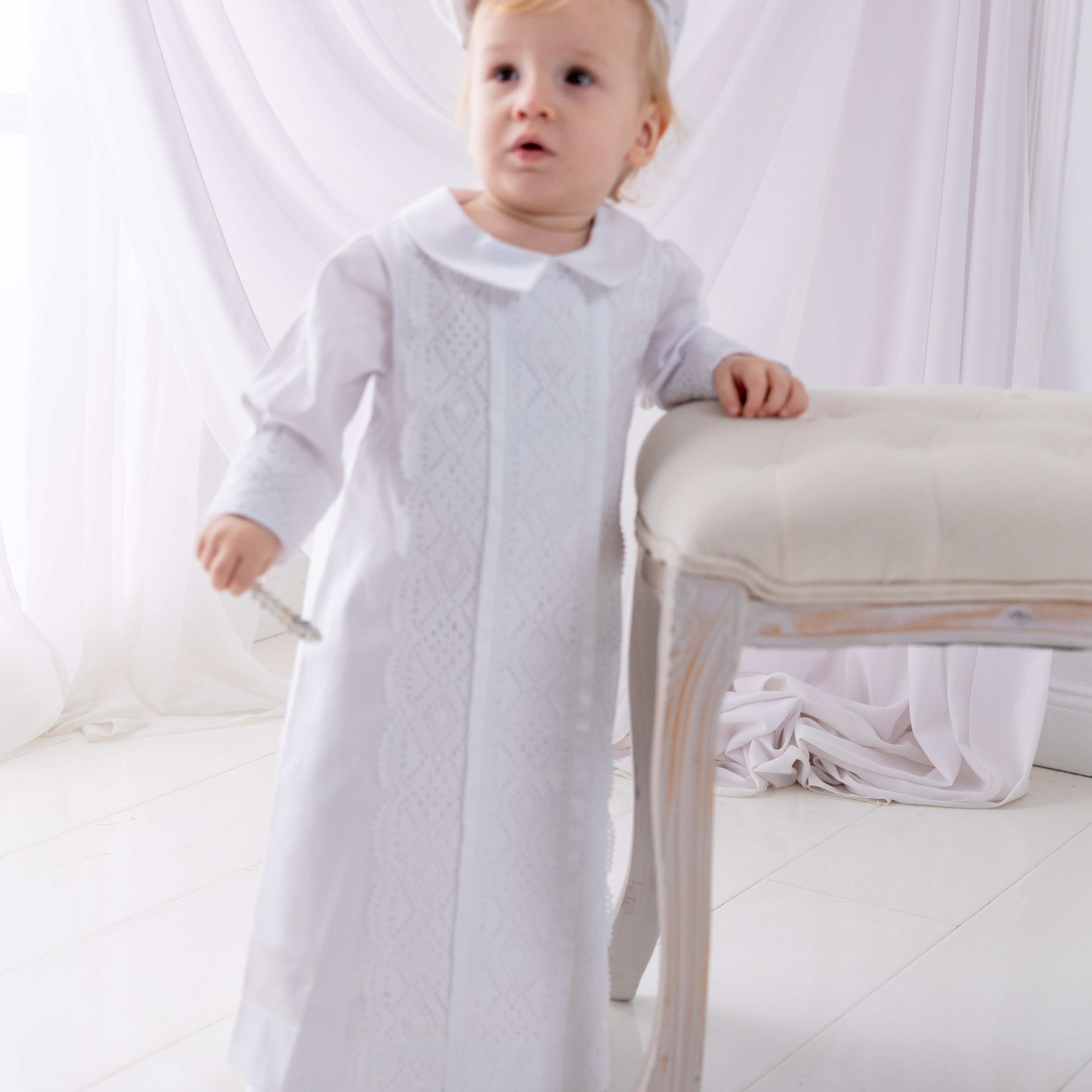 Baby in a white outfit standing on a white surface with a white curtain in the background