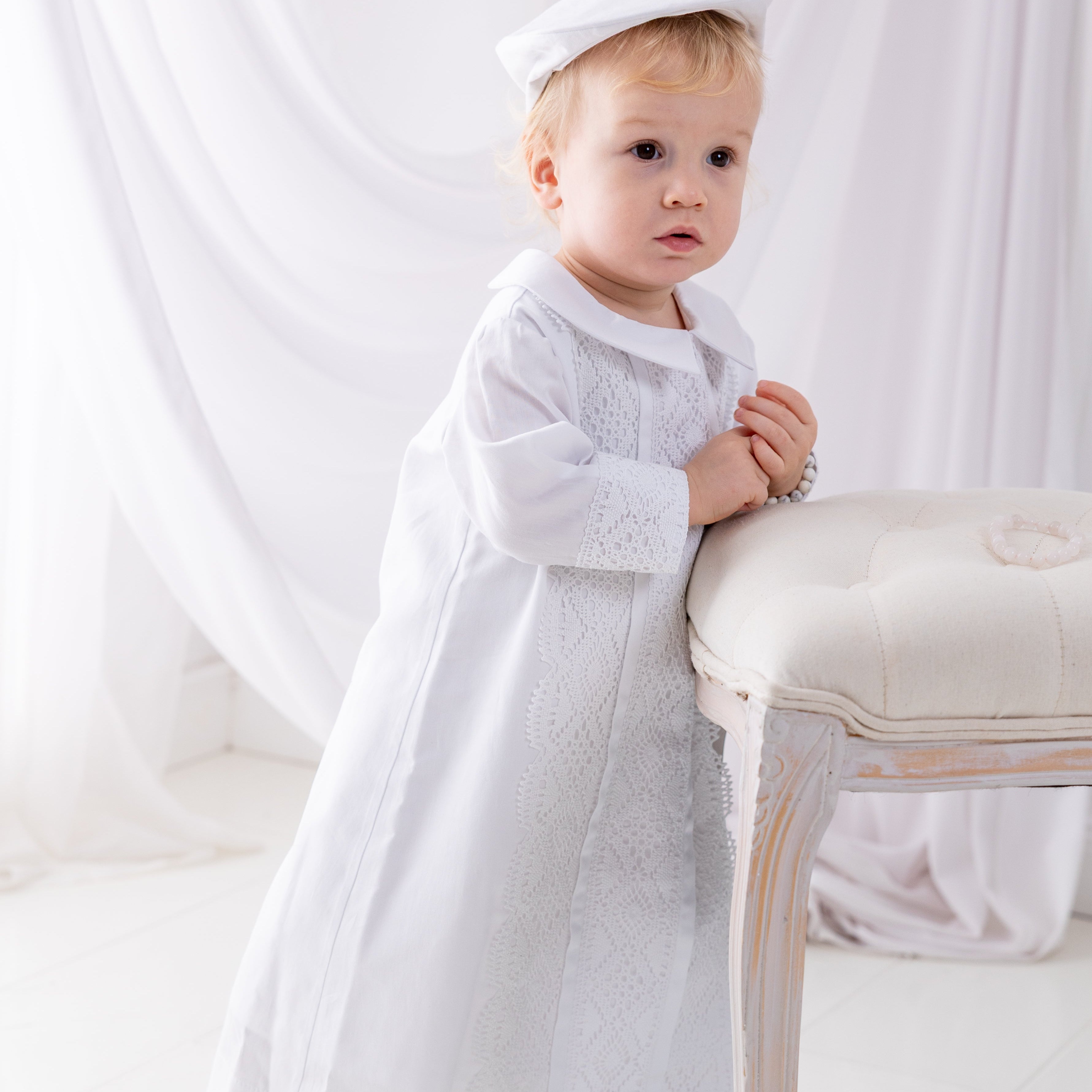 Child in a white outfit sitting on a chair against a white curtain background