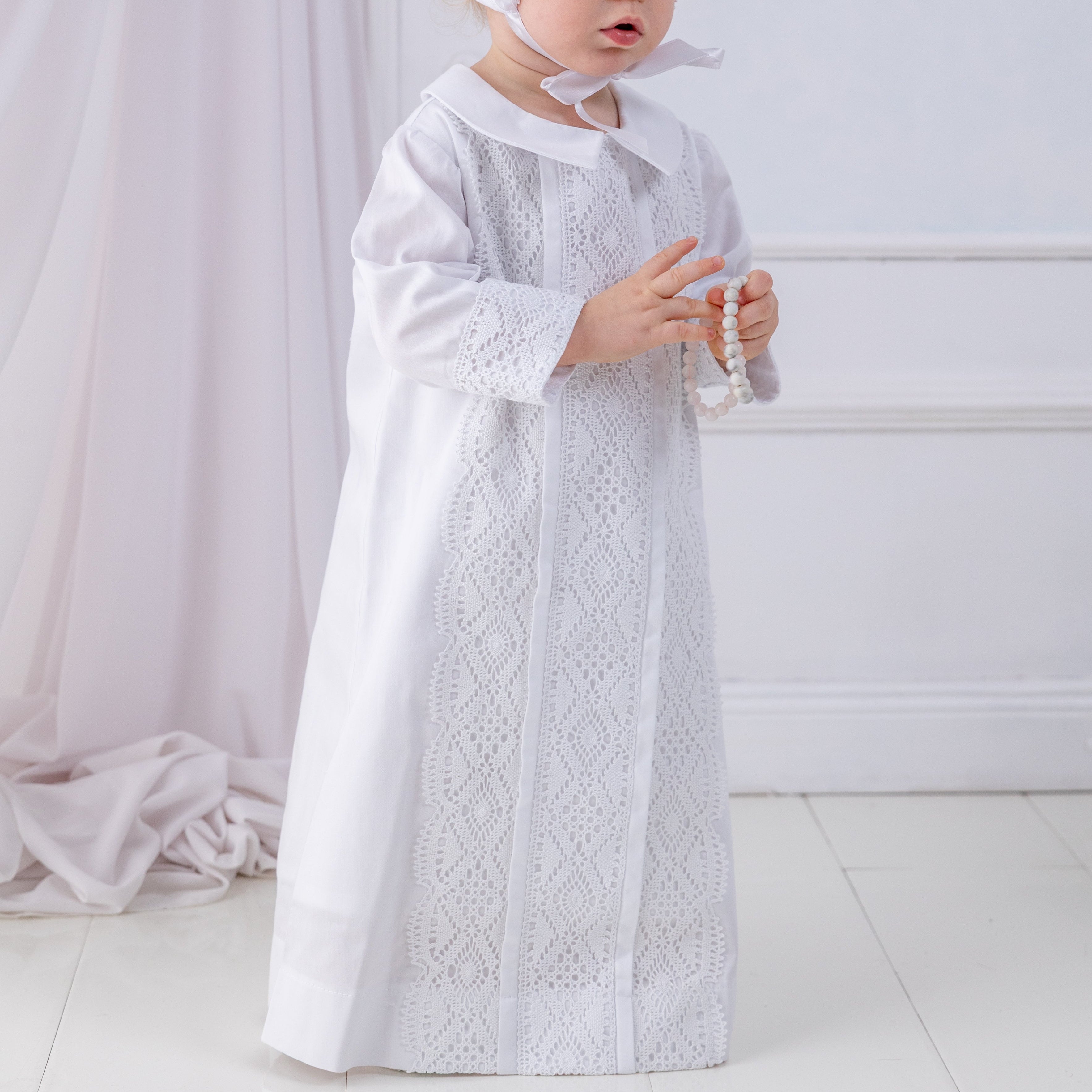 Child wearing a white lace baptism dress and bonnet in a softly lit room.