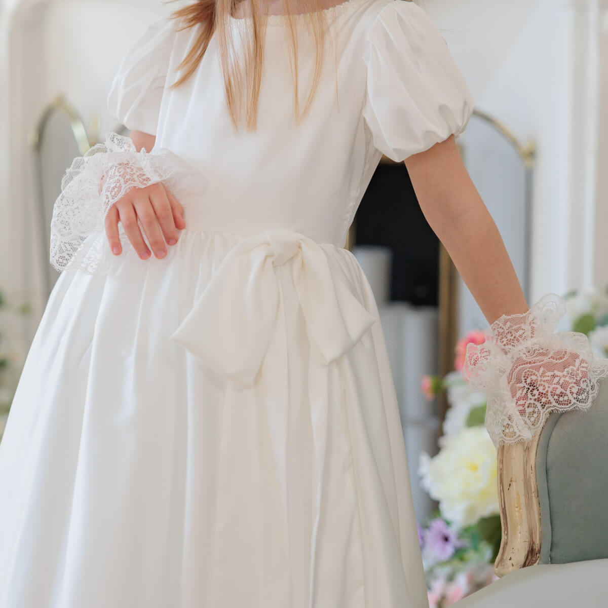 White dress with lace sleeves and cuffs worn by a person, blurred background with flowers.