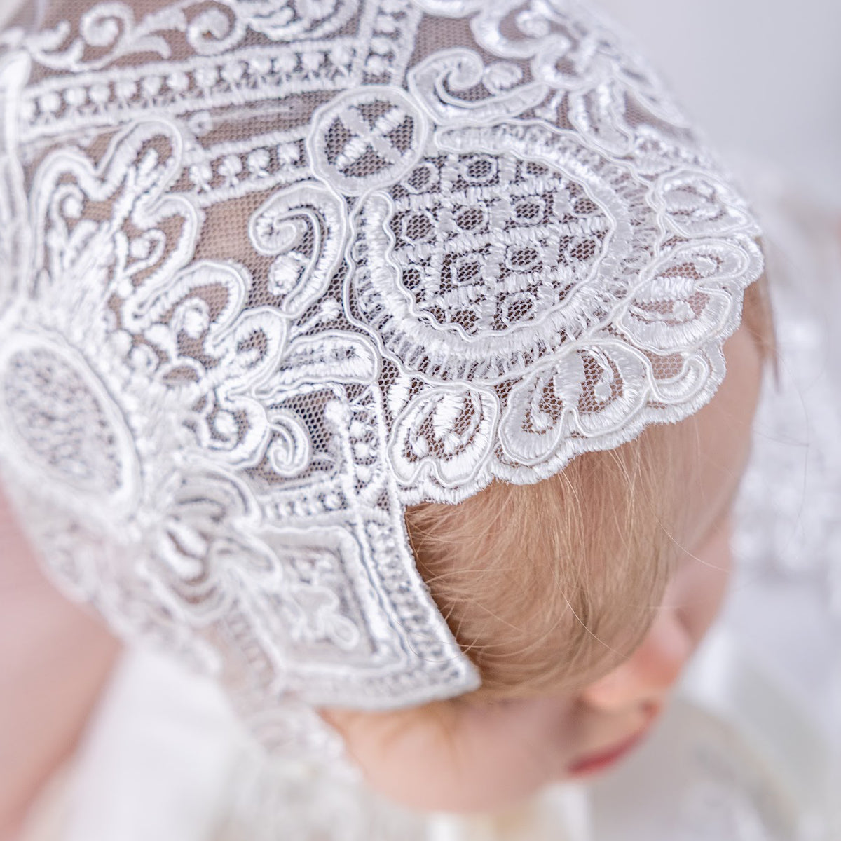 Baby in a lace baptism bonnet with a bonnet, sitting on a soft surface.