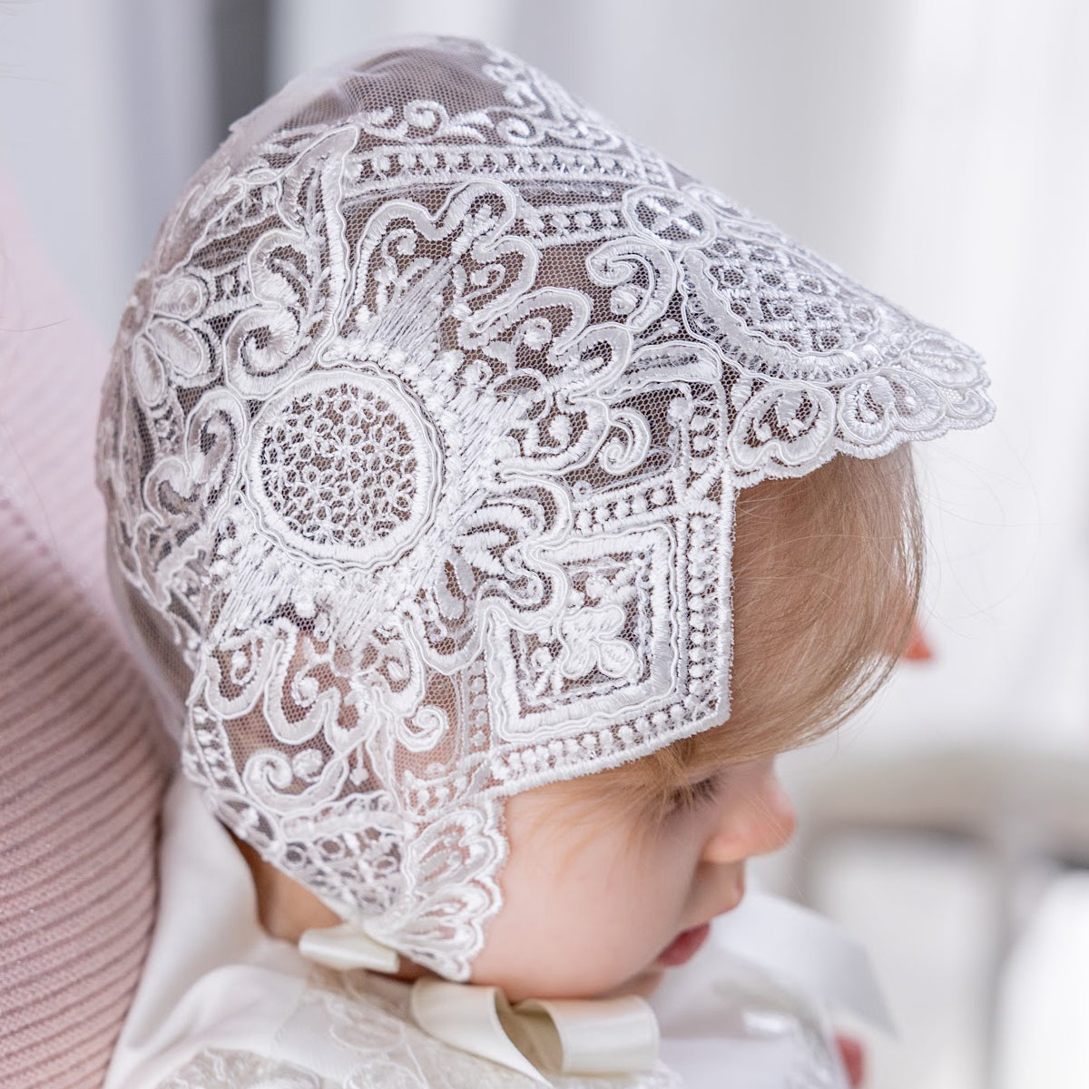 Baby in a lace baptism bonnet with a bonnet, sitting on a soft surface.