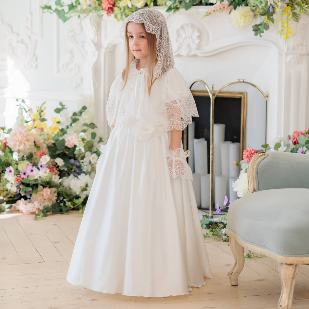Young girl in Silk First Communion Dress with Lace Mantilla standing in a decorated room with flowers and a mirror.