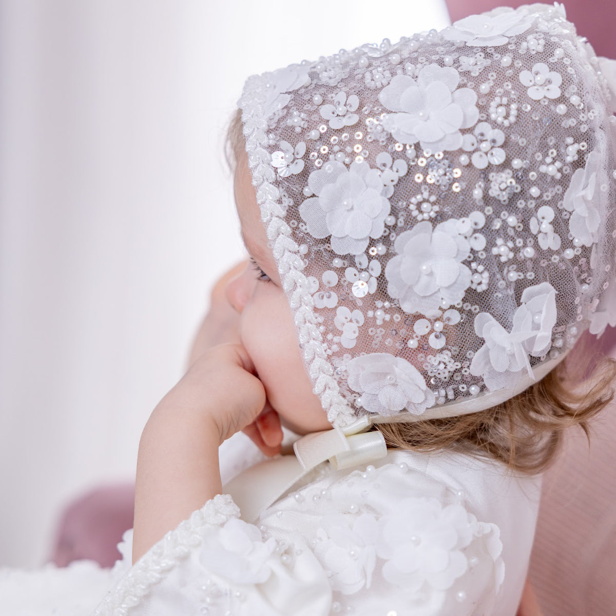 Child wearing a white lace bonnet with floral details and a bow, against a light background.
