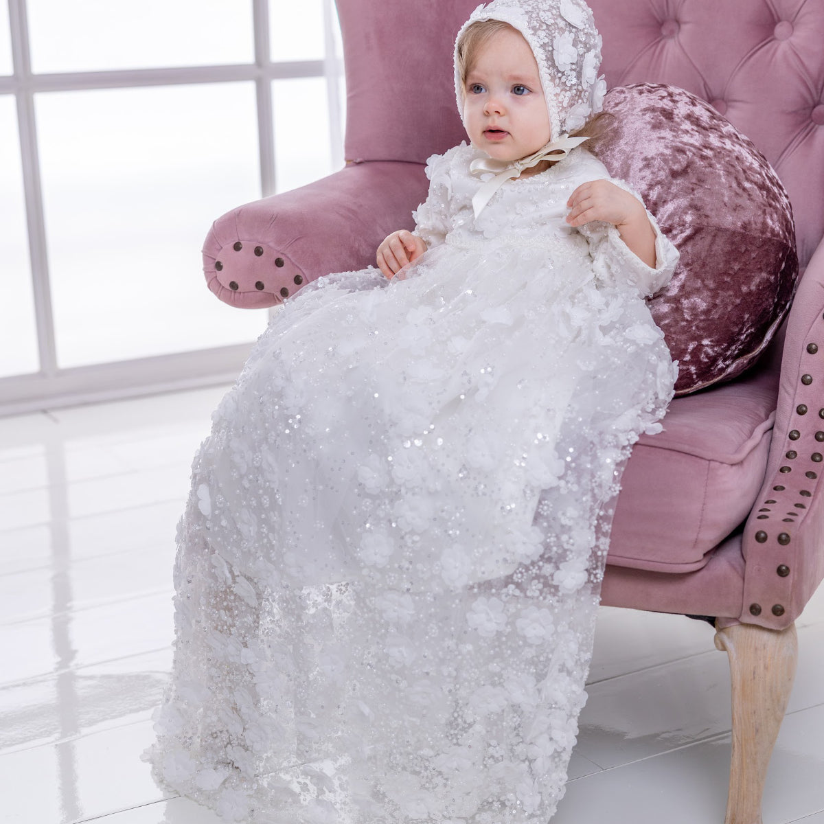 Baby in a white textured christening gown sitting on a pink chair.