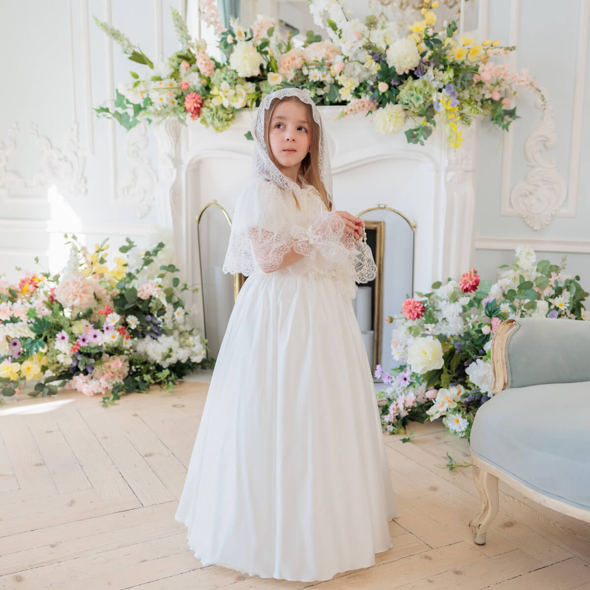 Young girl in Silk First Communion Dress with Lace Mantilla standing in a decorated room with flowers and furniture.