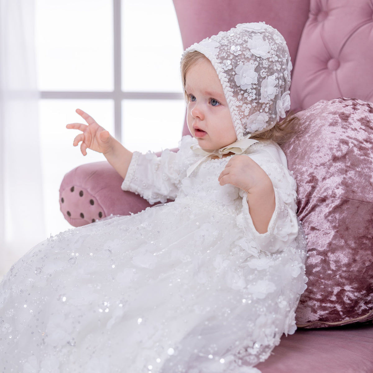 Baby in a white lace outfit sitting on a pink cushion