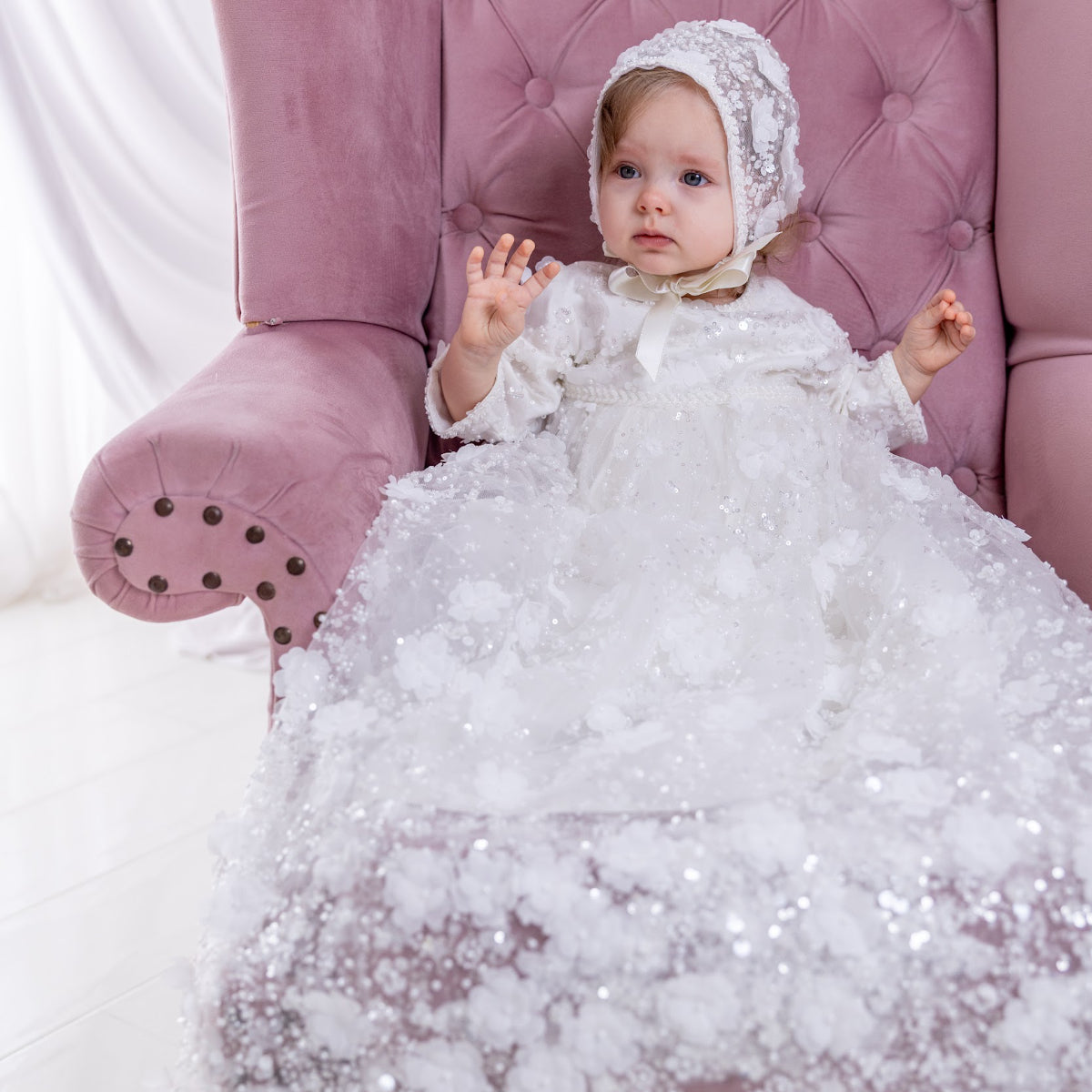 Baby in a white sparkly christening dress and bonnet sitting on a pink chair.