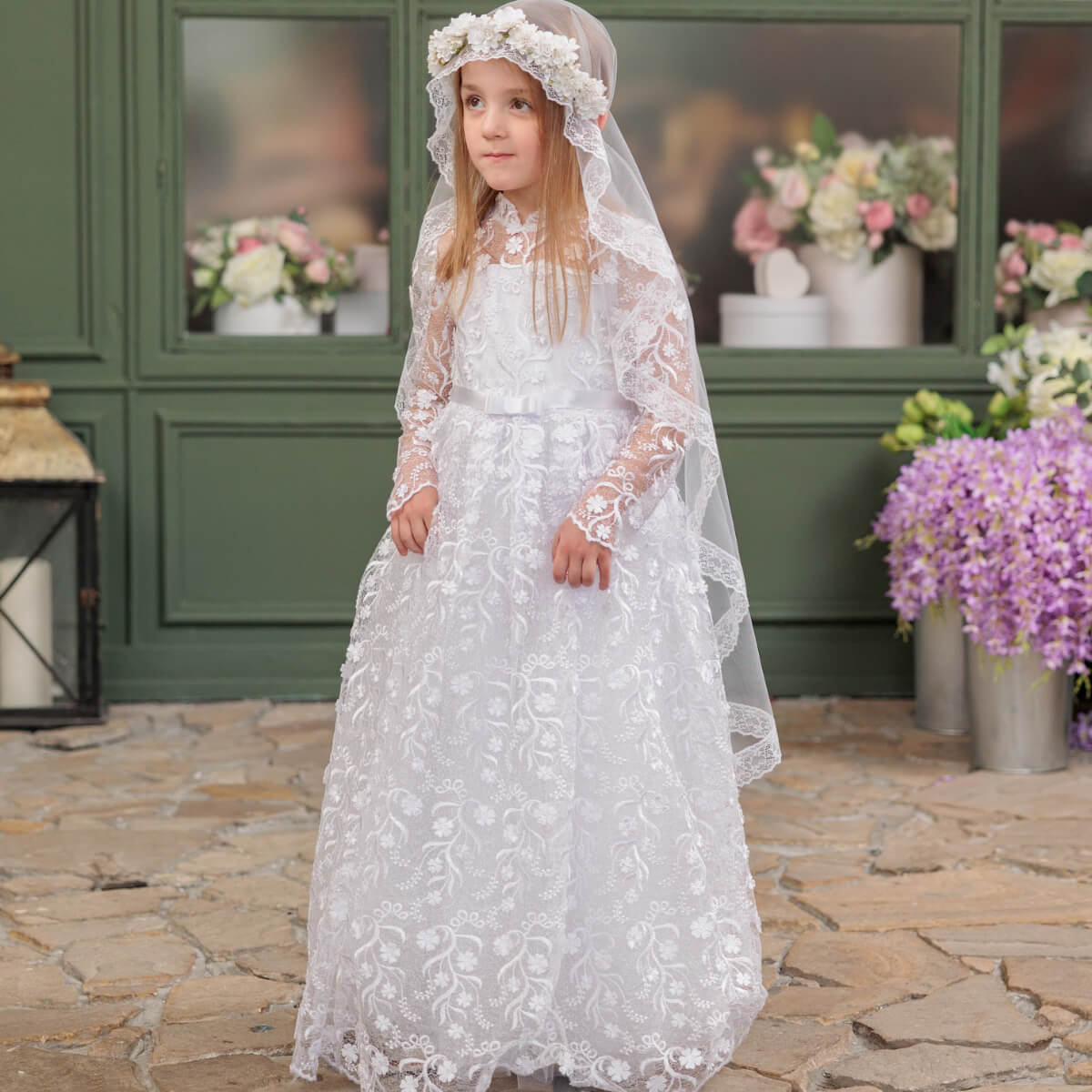 Young girl in a white lace communion dress standing outdoors with flowers and greenery in the background.
