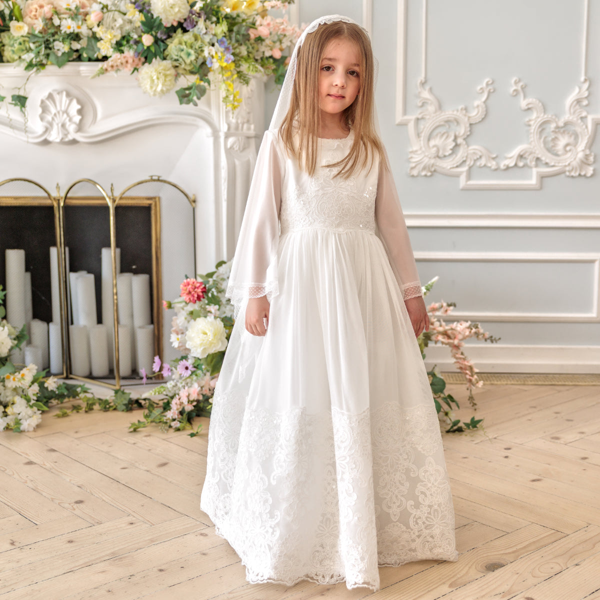 Young girl in Holy Communion Dress standing in a decorated room with flowers and a fireplace.