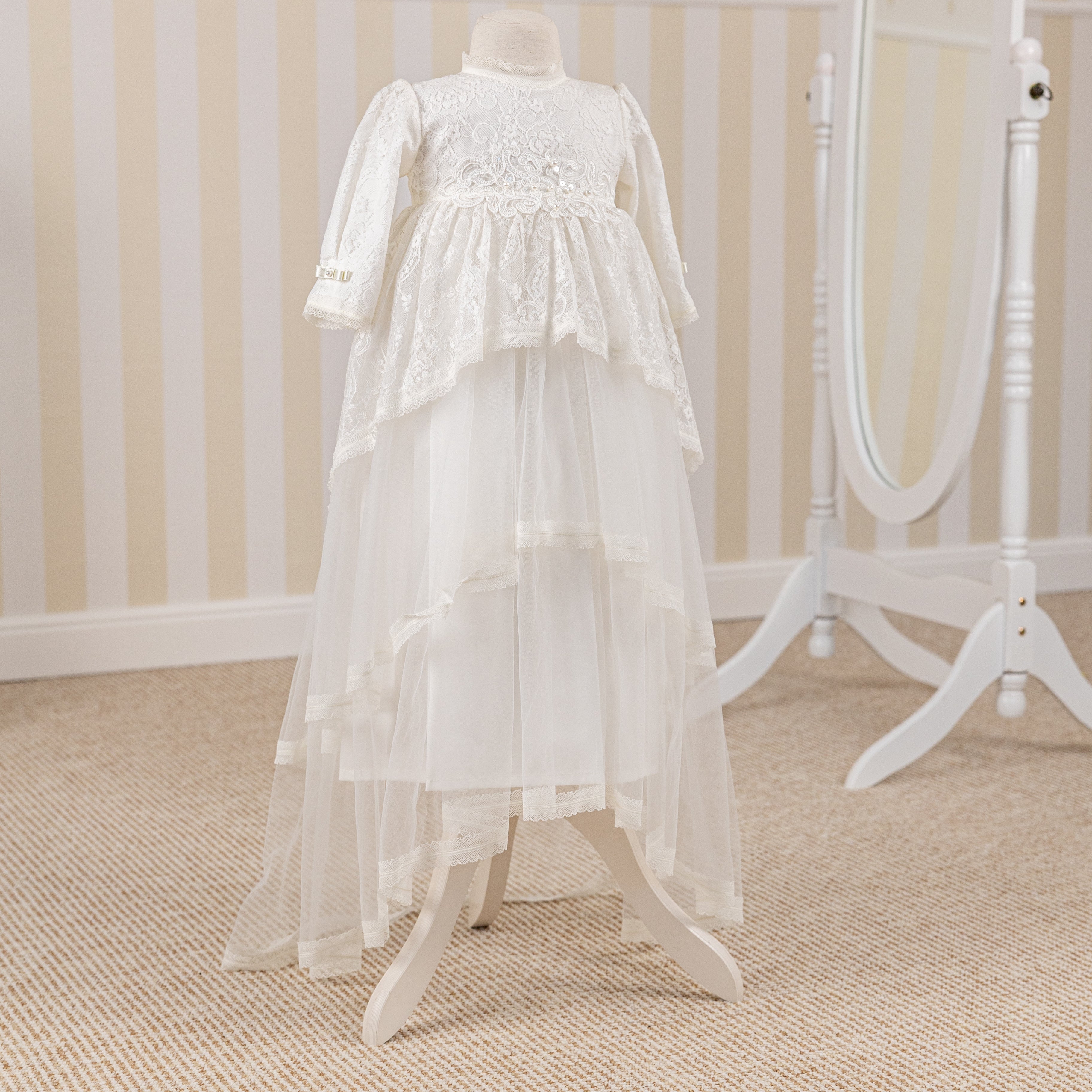 White lace baptism dress on a mannequin against a striped wall.