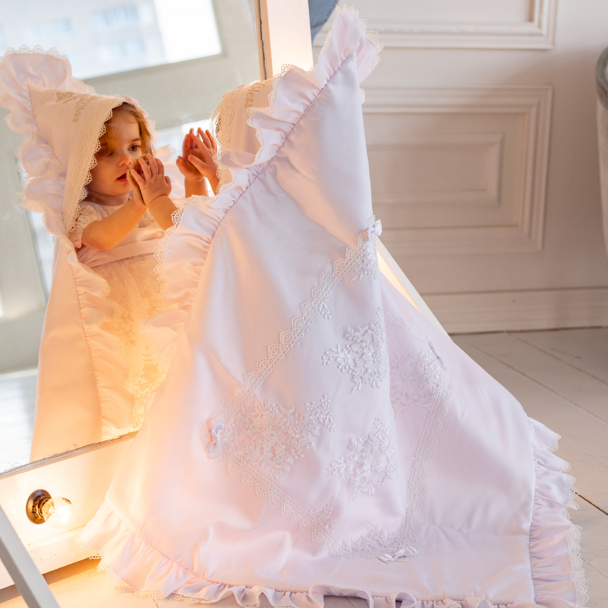 Child with white lace baptism blanket standing in a softly lit room.