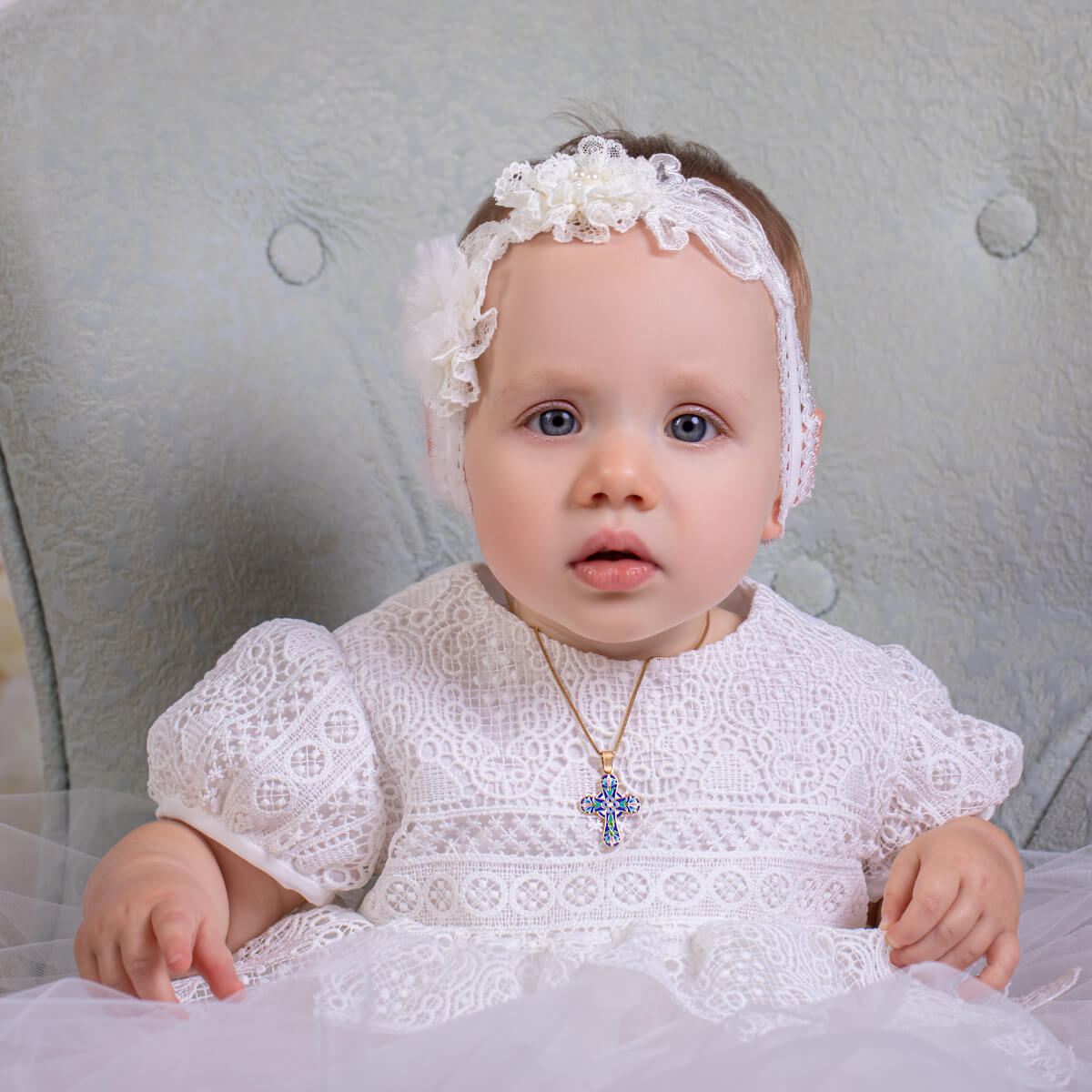 Baby in a white dress with a headband and necklace, sitting on a textured surface.