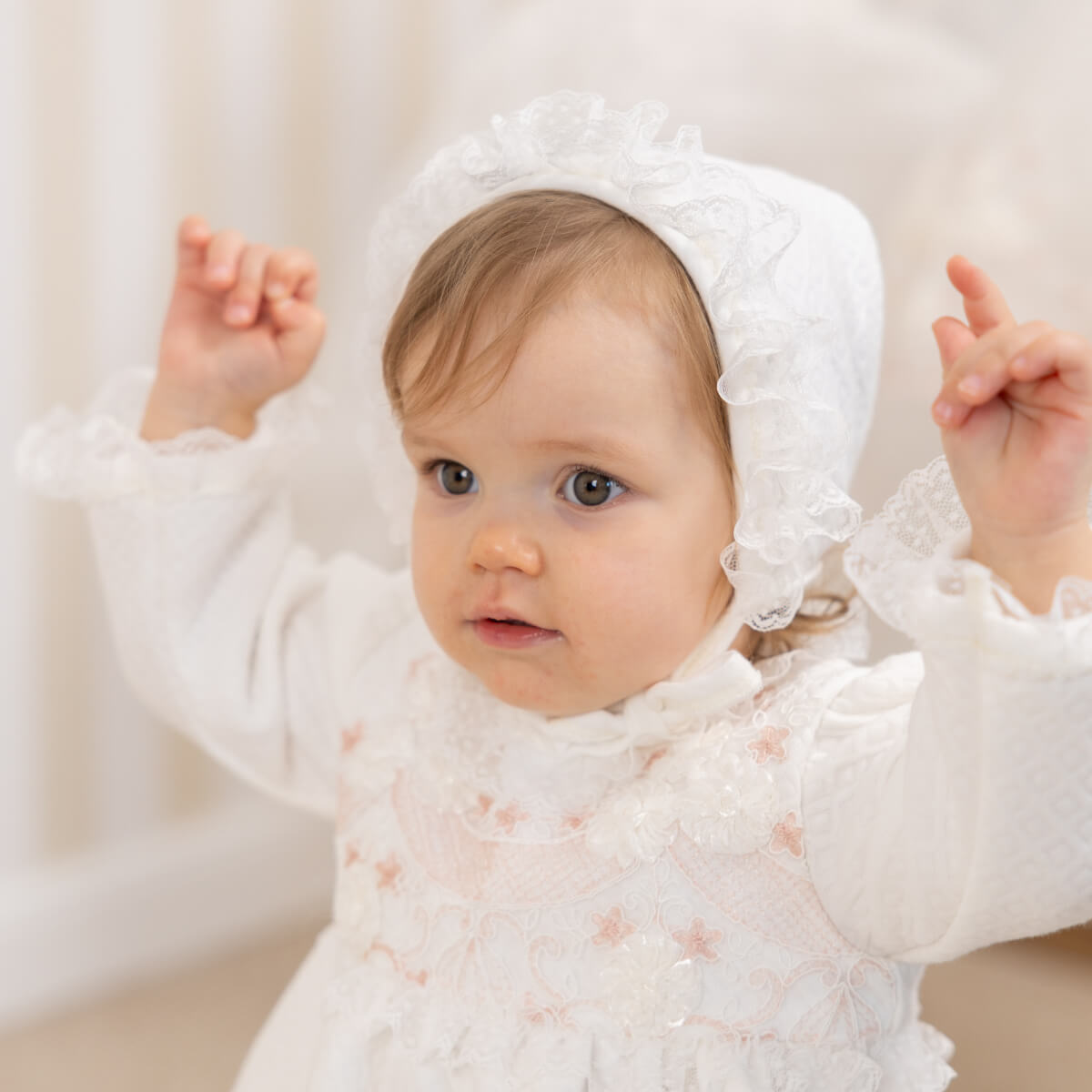 Baby in a white lace outfit with a Lace Baptism Bonnet against a neutral background