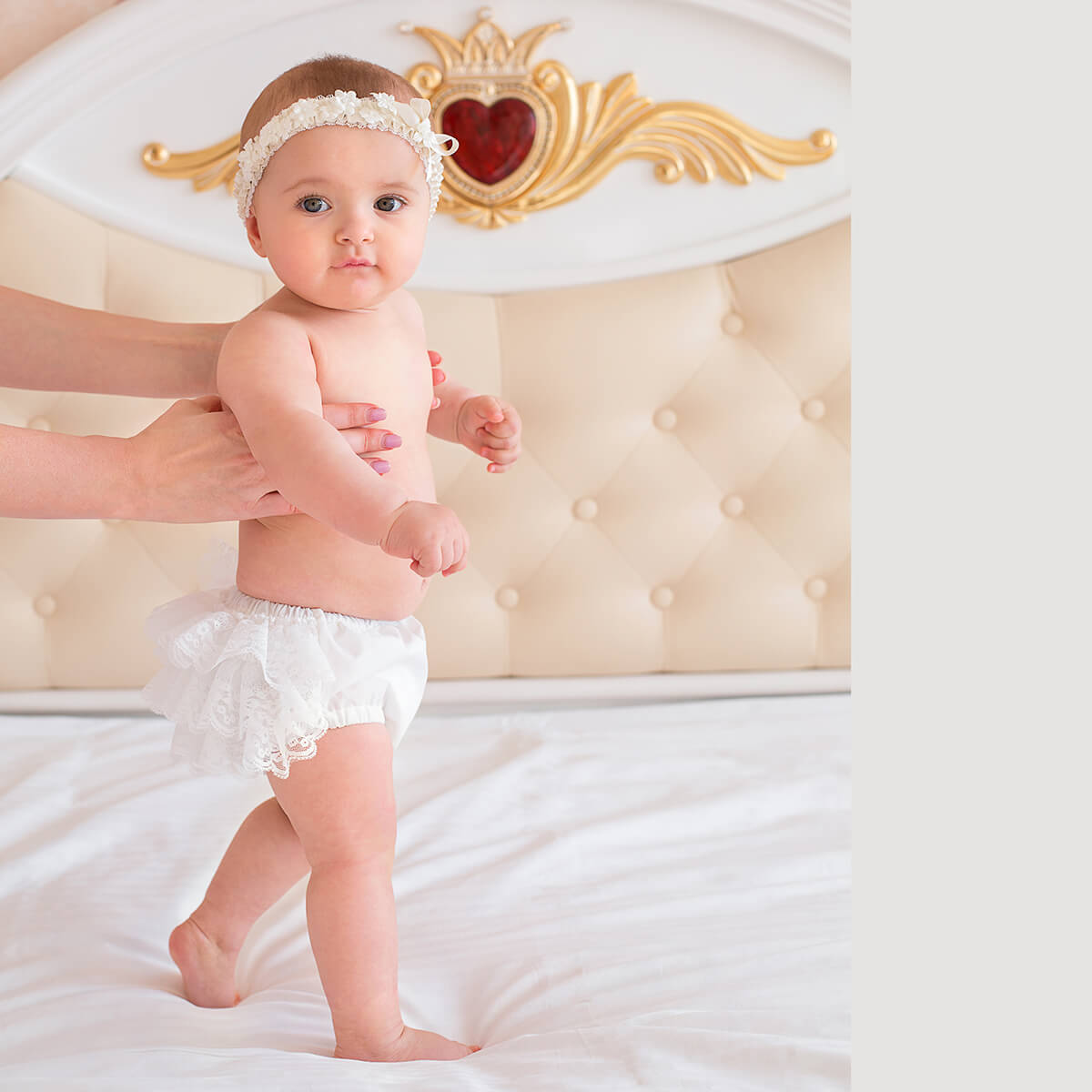 Baby in a Baby Keepsake Bloomersr and headband standing on a bed with a decorative headboard in the background.