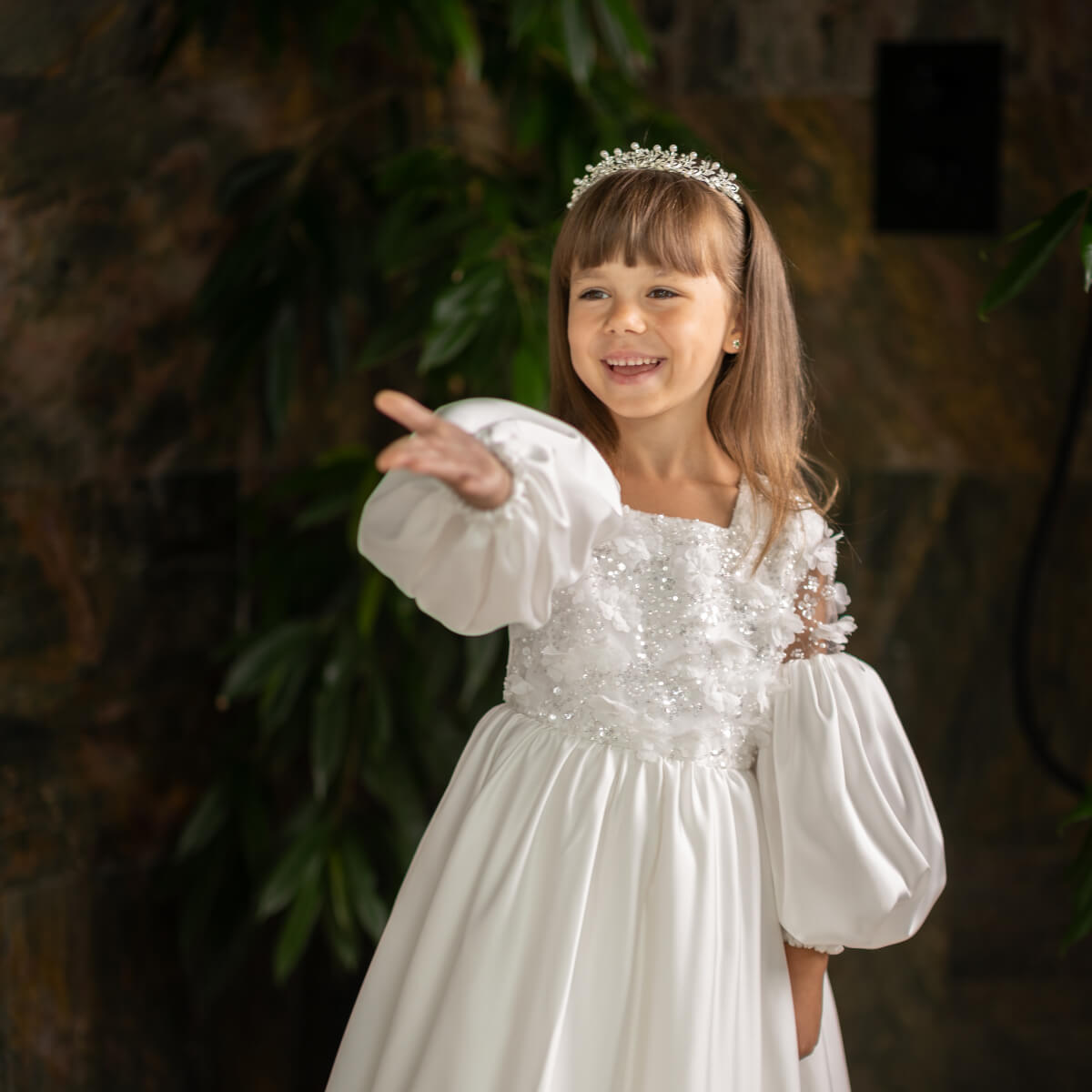 Young girl in a Silk First Communion Dress with puffed sleeves and a tiara, standing against a dark background.