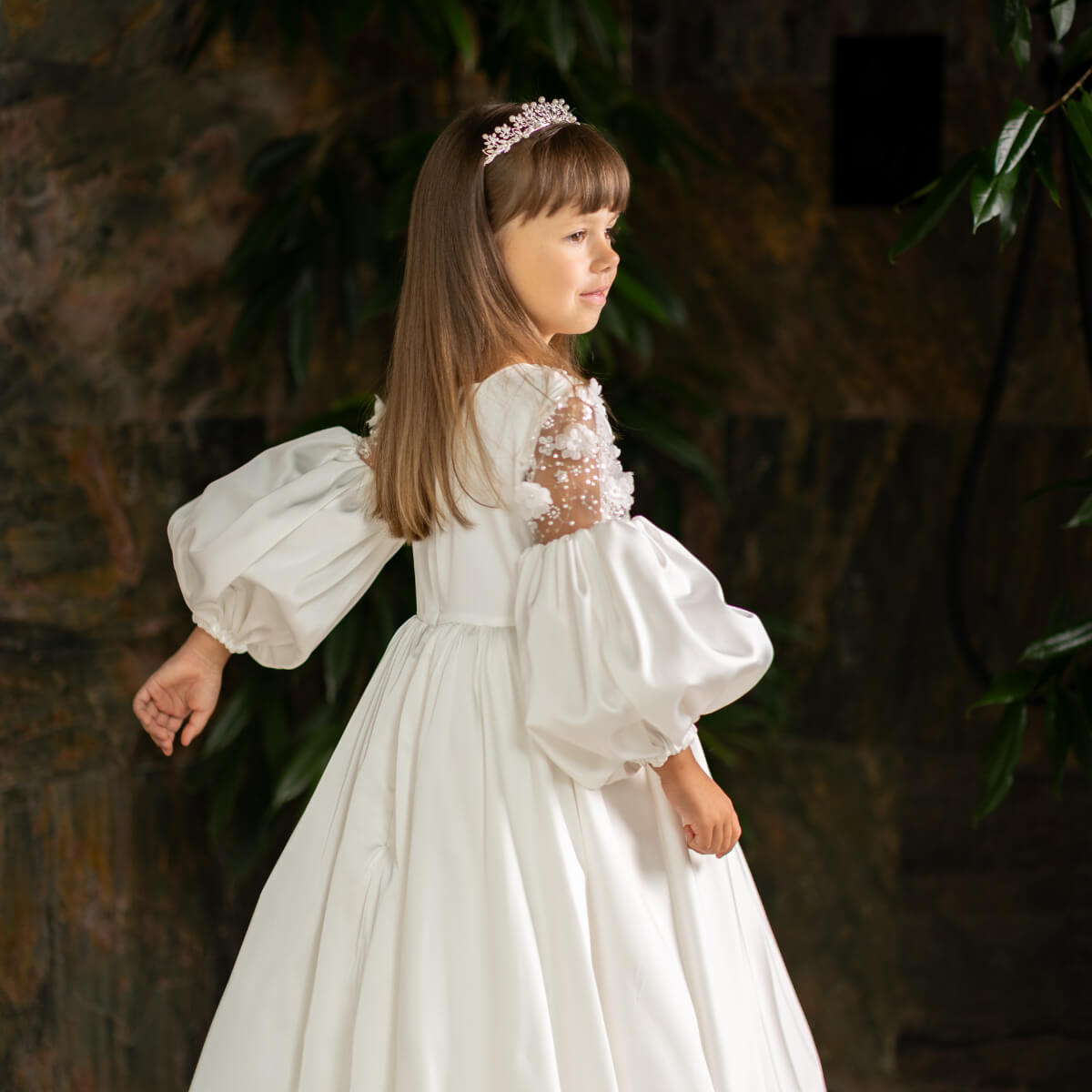 Young girl in a Silk First Communion Dress with puffed sleeves standing against a dark background.