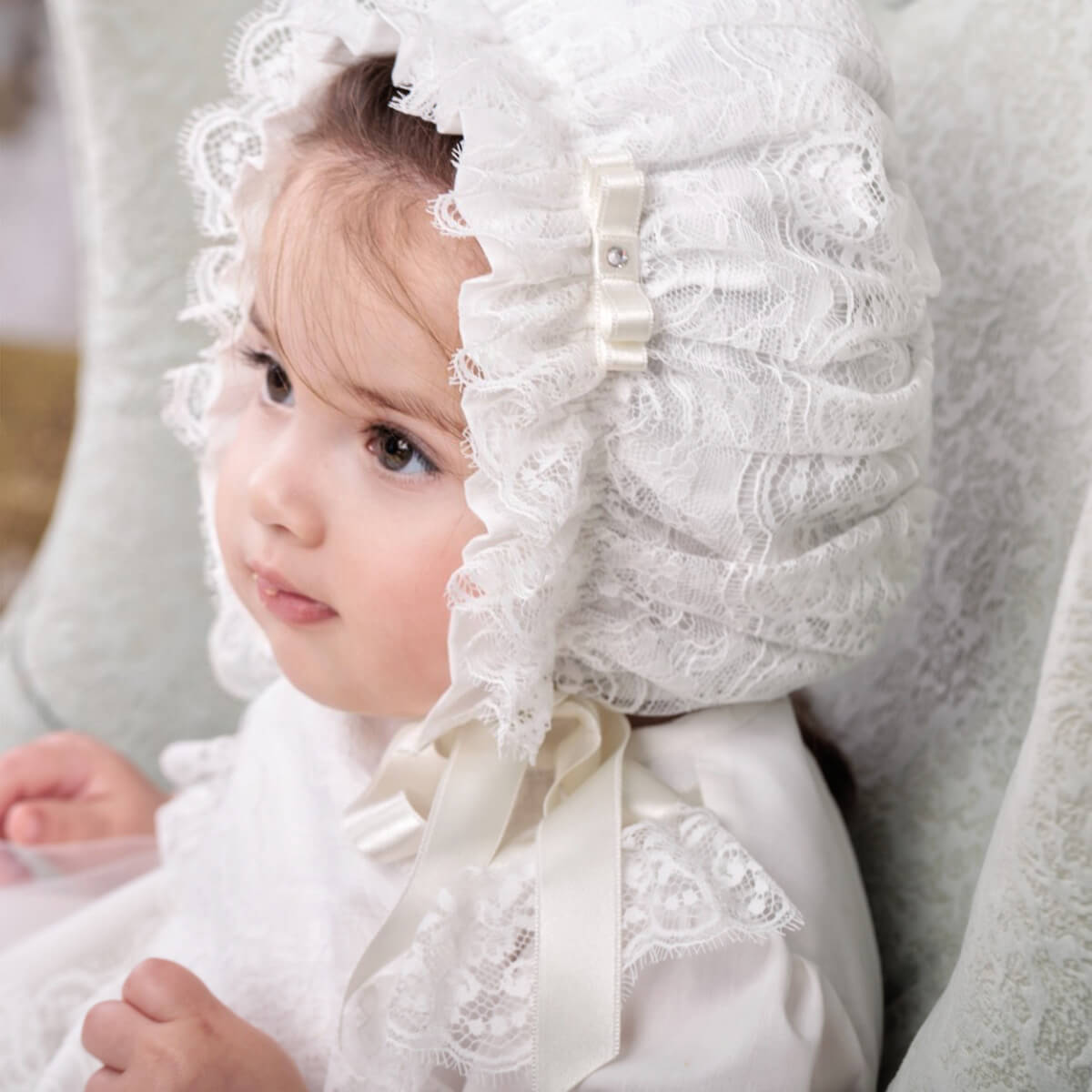 Child wearing a Lace Baptism Bonnet and dress, sitting on a soft surface.
