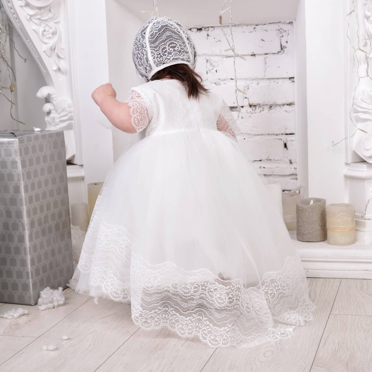 Child in a Baptism Dress with Train and bonnet in a decorated room with white walls and decorative elements.