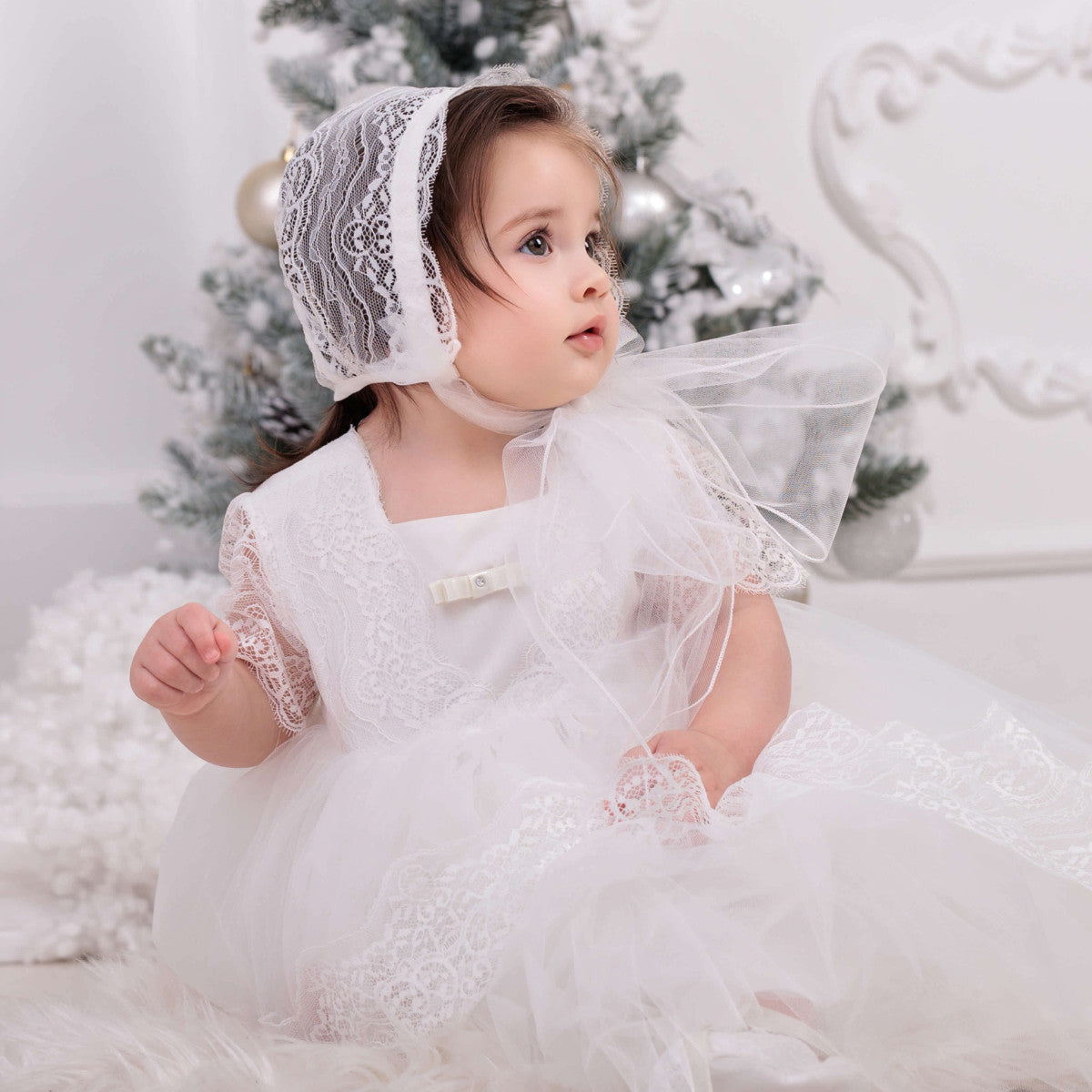 Child in a Baptism Dress with Train and bonnet sitting in front of a decorated Christmas tree.