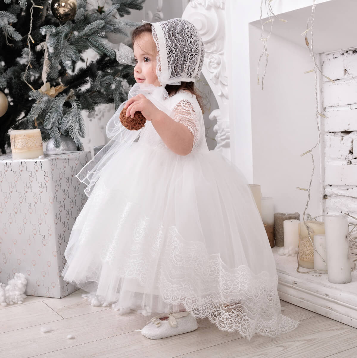 Child in a Baptism Dress with Train and bonnet standing in a decorated room with Christmas trees and lights.