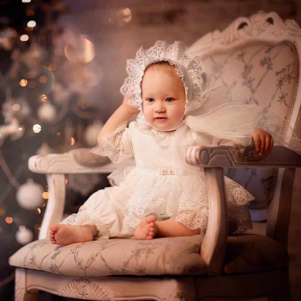 Baby in a white lace outfit sitting on an ornate chair with a blurred Christmas tree in the background.