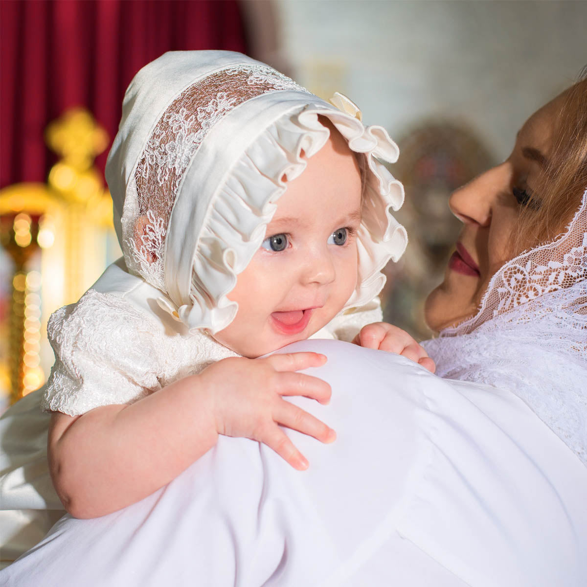 Baby in a Silk Christening Bonnet being held by a woman in a similar outfit, with a blurred background.