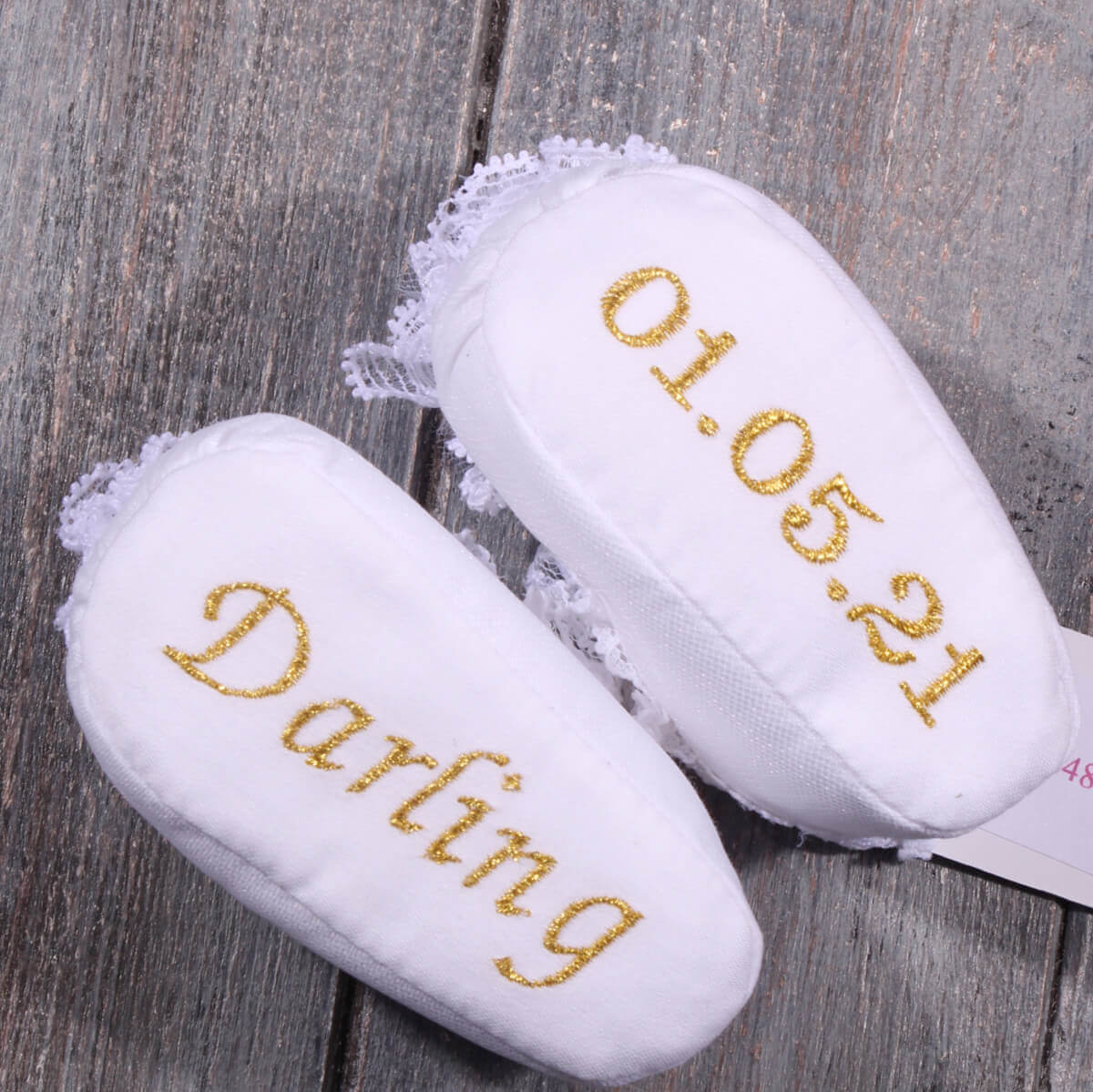 White baby shoes with gold text 'Darling' and '01.05.21' on a wooden surface