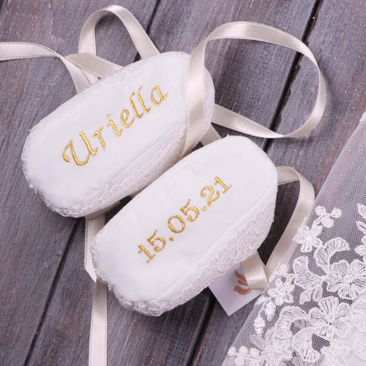 White baptism booties with embroidered names and dates on a wooden surface with lace fabric.